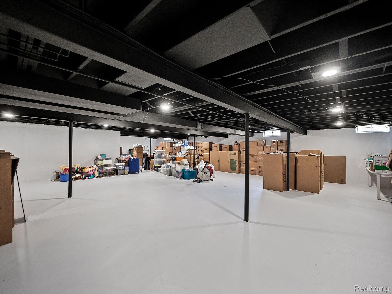 This is a spacious basement featuring a painted white floor and walls, contrasted by a black open ceiling with exposed beams and ductwork. The area is filled with numerous cardboard boxes and storage containers, suggesting ample storage space. The room is well-lit with recessed lighting, and support beams are strategically placed throughout the area.