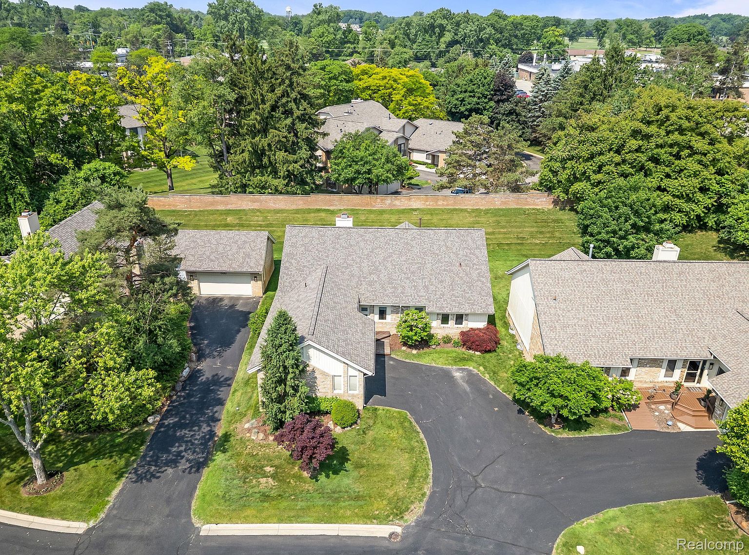 This aerial shot showcases a well-maintained residential property with a gray roof, lush green lawns, and mature trees. A paved driveway leads to the house and a detached garage. The property is surrounded by a brick wall and other homes, creating a sense of community.