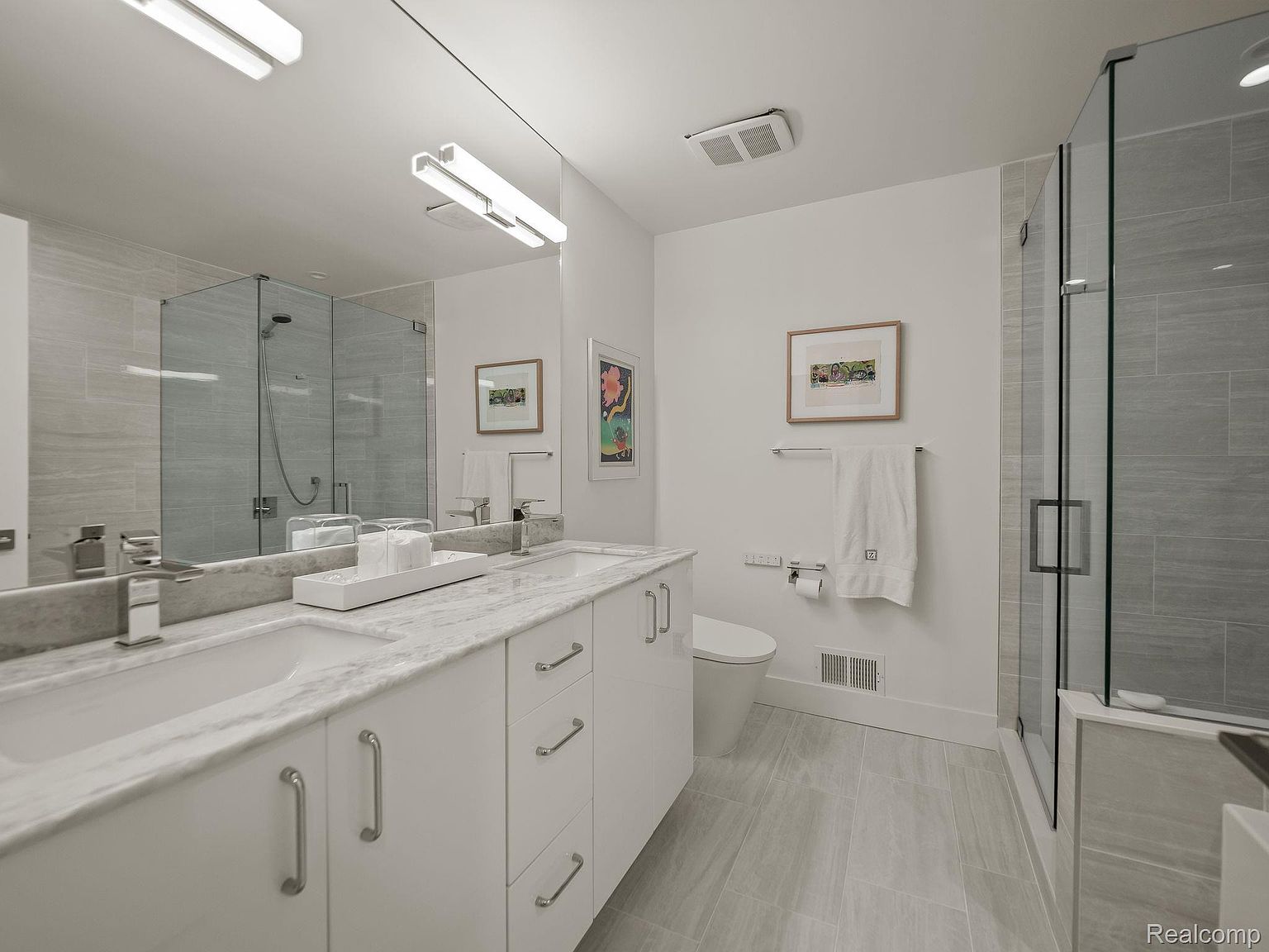 This is a well-lit primary bathroom featuring a double vanity with a white marble countertop and white cabinetry. A large mirror spans the length of the vanity, reflecting the shower enclosure with glass doors. The bathroom has a modern and clean aesthetic with gray tile flooring and walls, creating a spa-like atmosphere.