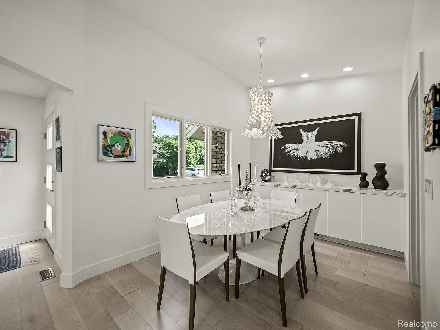 This is an interior shot of a modern dining room. The room features an oval marble table surrounded by white chairs with dark wood legs, a unique floral chandelier, and a large black and white art piece. A built-in white cabinet provides additional storage, and the light wood flooring adds warmth to the space.