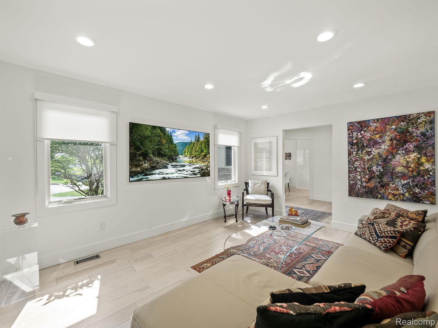 This is a bright and airy living room featuring white walls, light wood flooring, and recessed lighting. A large television displays a nature scene, and a colorful abstract painting hangs on the wall. The room is furnished with a beige sofa, patterned pillows, a glass coffee table, and an accent chair, creating a comfortable and stylish living space.