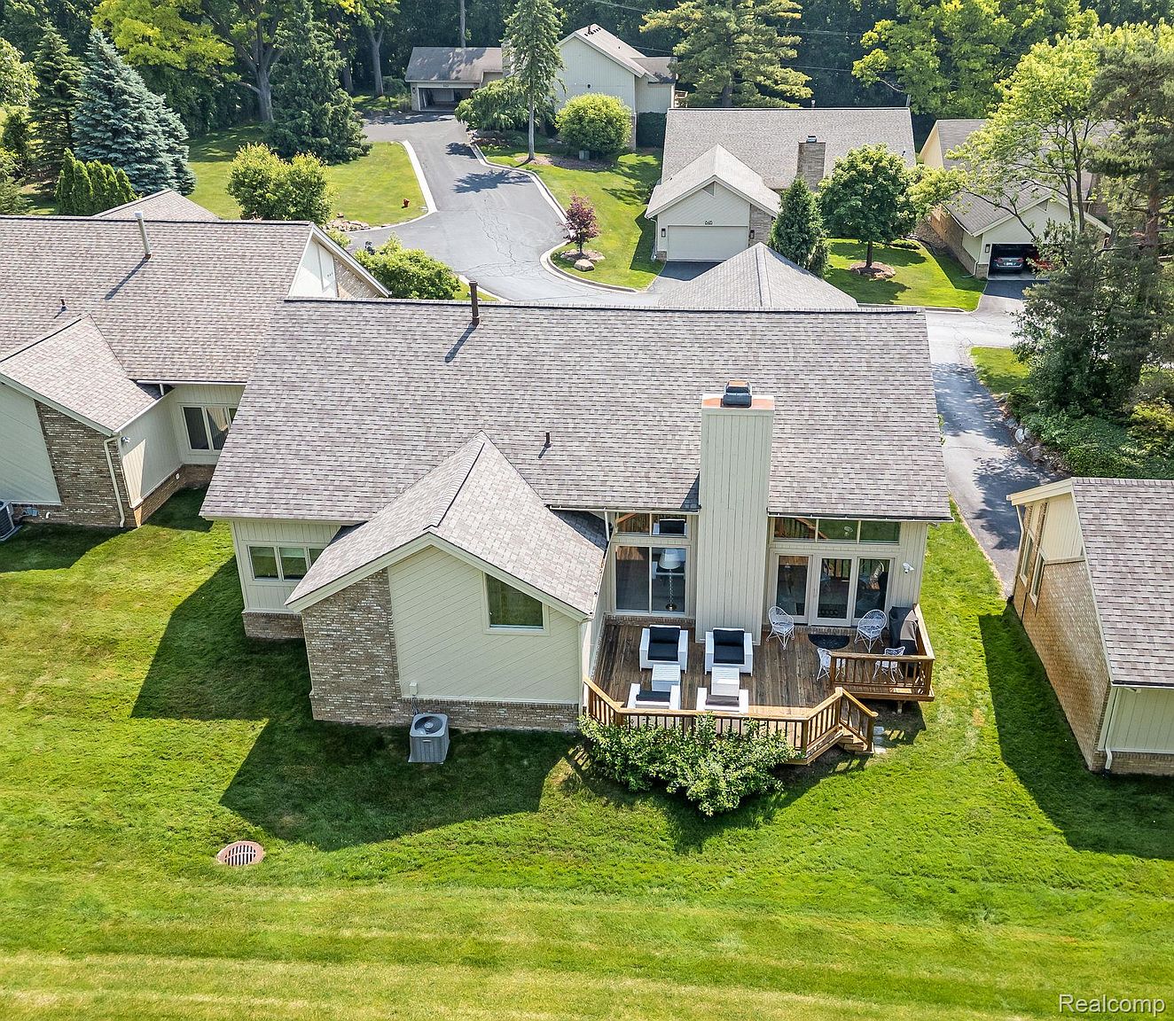 This aerial shot showcases a well-maintained residential property with lush green lawns and mature trees. The house features a neutral-toned exterior with a prominent chimney and a wooden deck furnished with outdoor seating. The surrounding neighborhood appears quiet and upscale, with paved roads and well-spaced homes.