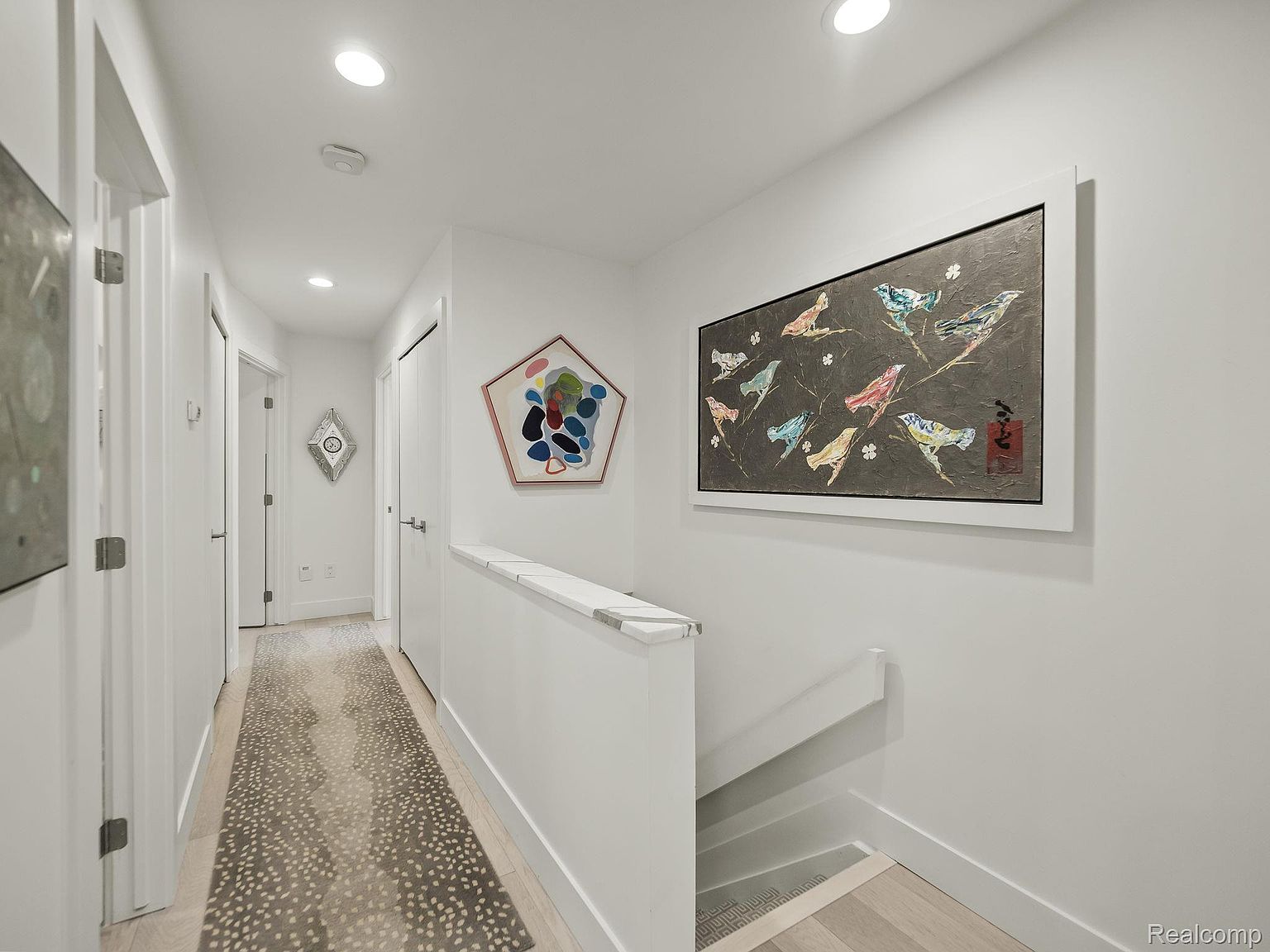This is a well-lit hallway featuring white walls, trim, and doors, creating a clean and modern aesthetic. Artwork adorns the walls, adding visual interest, while a patterned runner rug adds warmth and texture to the light wood flooring. The hallway leads to a staircase, suggesting a multi-level home.