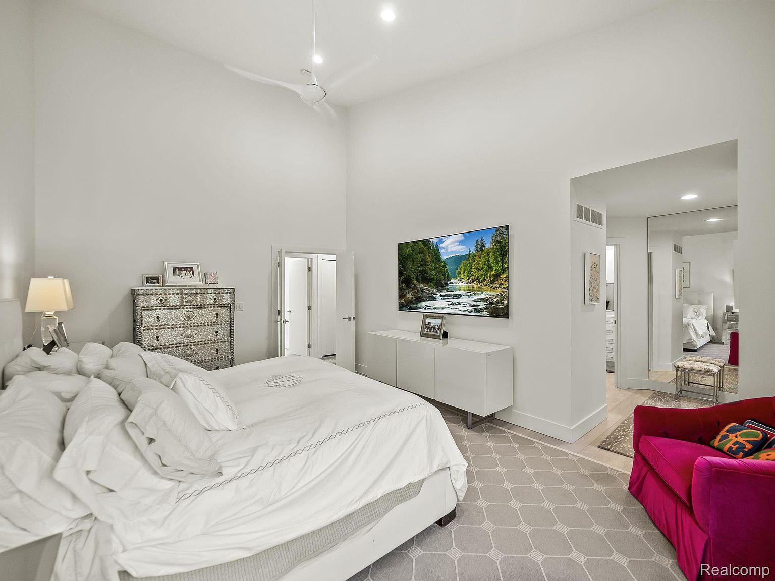 This is a bright and airy primary bedroom featuring a large bed with white linens, a decorative chest of drawers, and a wall-mounted television. The room has high ceilings and a neutral color palette, creating a serene and spacious atmosphere. A glimpse into an adjacent room reveals a walk-in closet and additional living space, enhancing the suite's appeal.