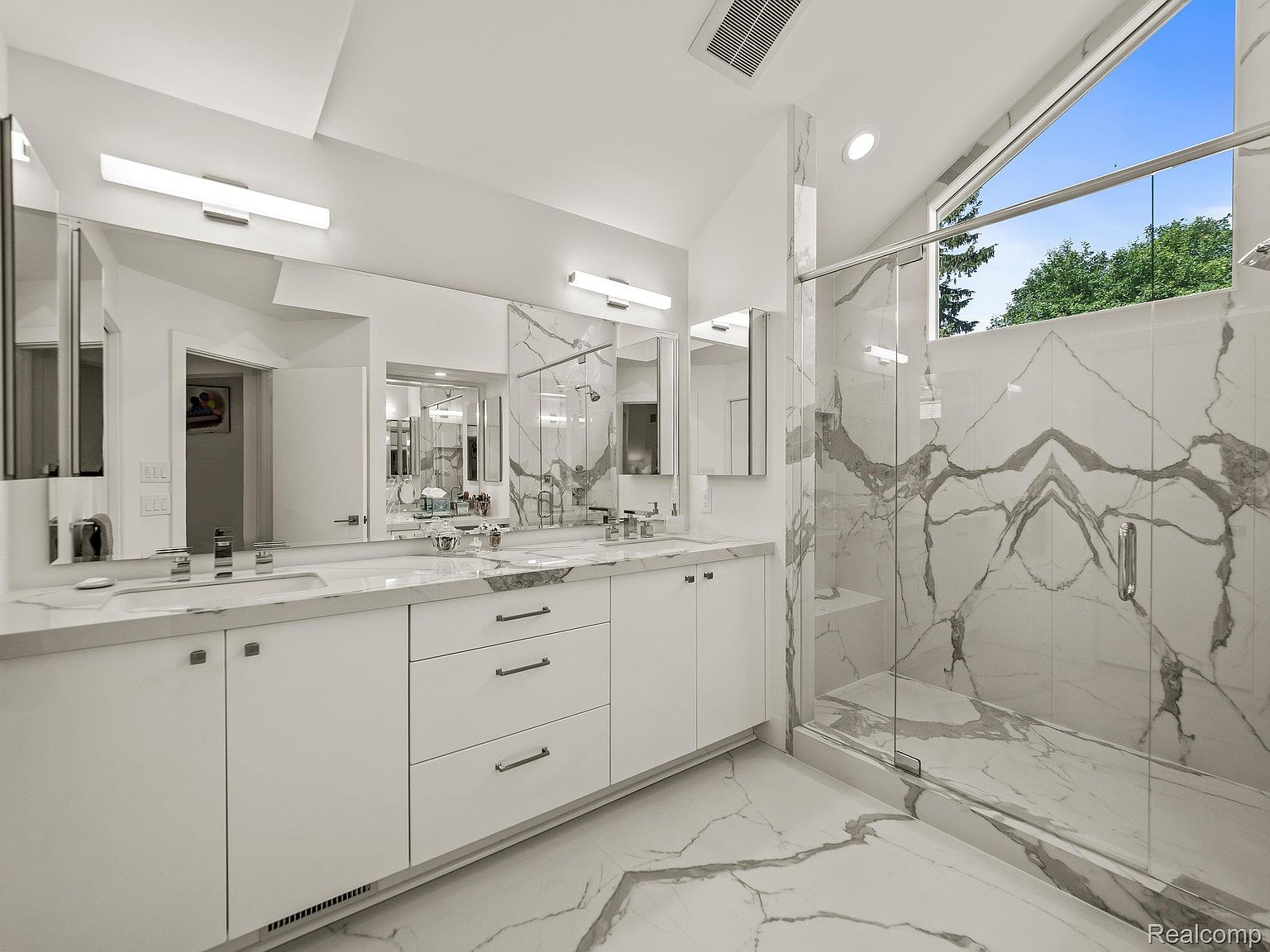 This is a bright and modern primary bathroom featuring a double vanity with white cabinetry and marble countertops. A large, glass-enclosed shower with marble walls adds a luxurious touch. The room is well-lit with contemporary light fixtures and natural light from a large window, creating a clean and spa-like atmosphere.