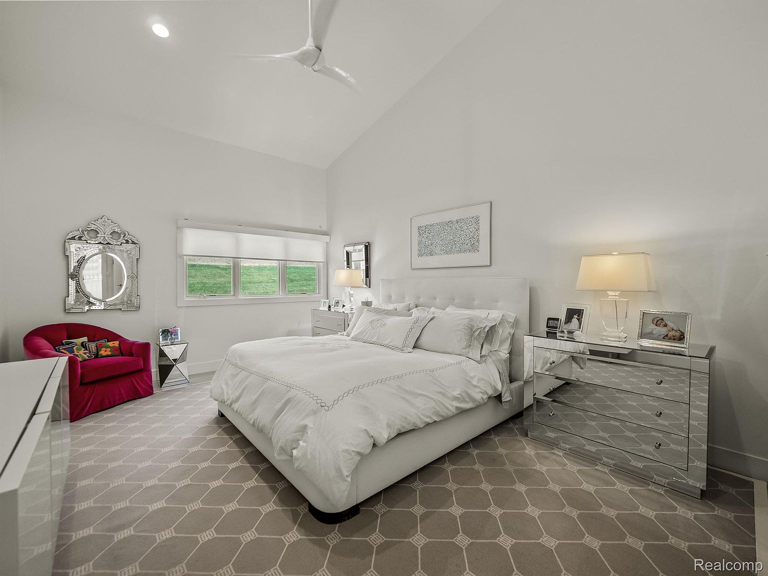 This is a bright and airy primary bedroom featuring a large bed with white linens, mirrored nightstands, and a decorative mirror. The room has a neutral color palette with a patterned carpet and a vaulted ceiling, creating a serene and luxurious atmosphere. A red chair adds a pop of color to the space.