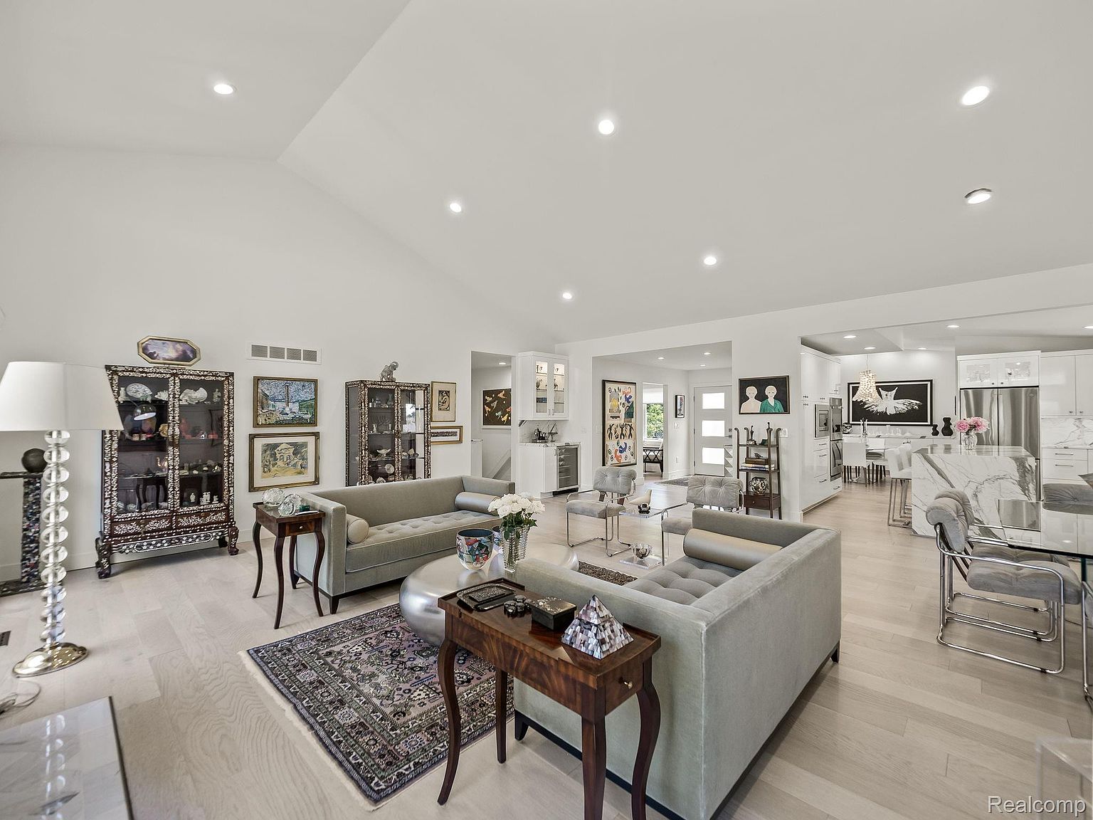 This is an interior shot of a spacious living room featuring two light gray sofas, wooden side tables, and a decorative rug. The room is well-lit with recessed lighting and natural light, showcasing a modern and elegant design. Display cabinets filled with decorative items add character to the space, which seamlessly transitions into an open-concept kitchen and dining area.