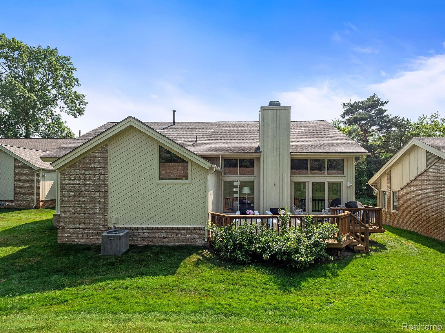 This is a rear view of a residential property, showcasing a well-maintained lawn and a wooden deck. The house features a combination of brick and siding, with large windows offering ample natural light. A chimney rises prominently from the roof, adding to the architectural interest.