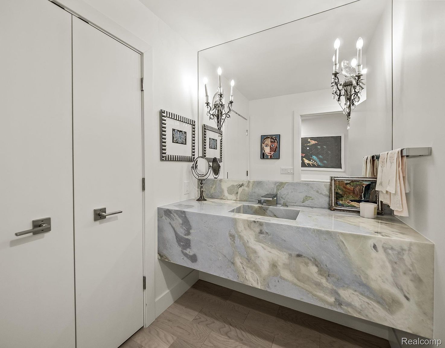 This is a well-lit bathroom featuring a modern floating vanity with a marble countertop and integrated sink. A large mirror spans the wall above the vanity, reflecting decorative sconces and artwork. The room has a clean, minimalist aesthetic with white walls and light wood flooring, creating a bright and airy atmosphere.