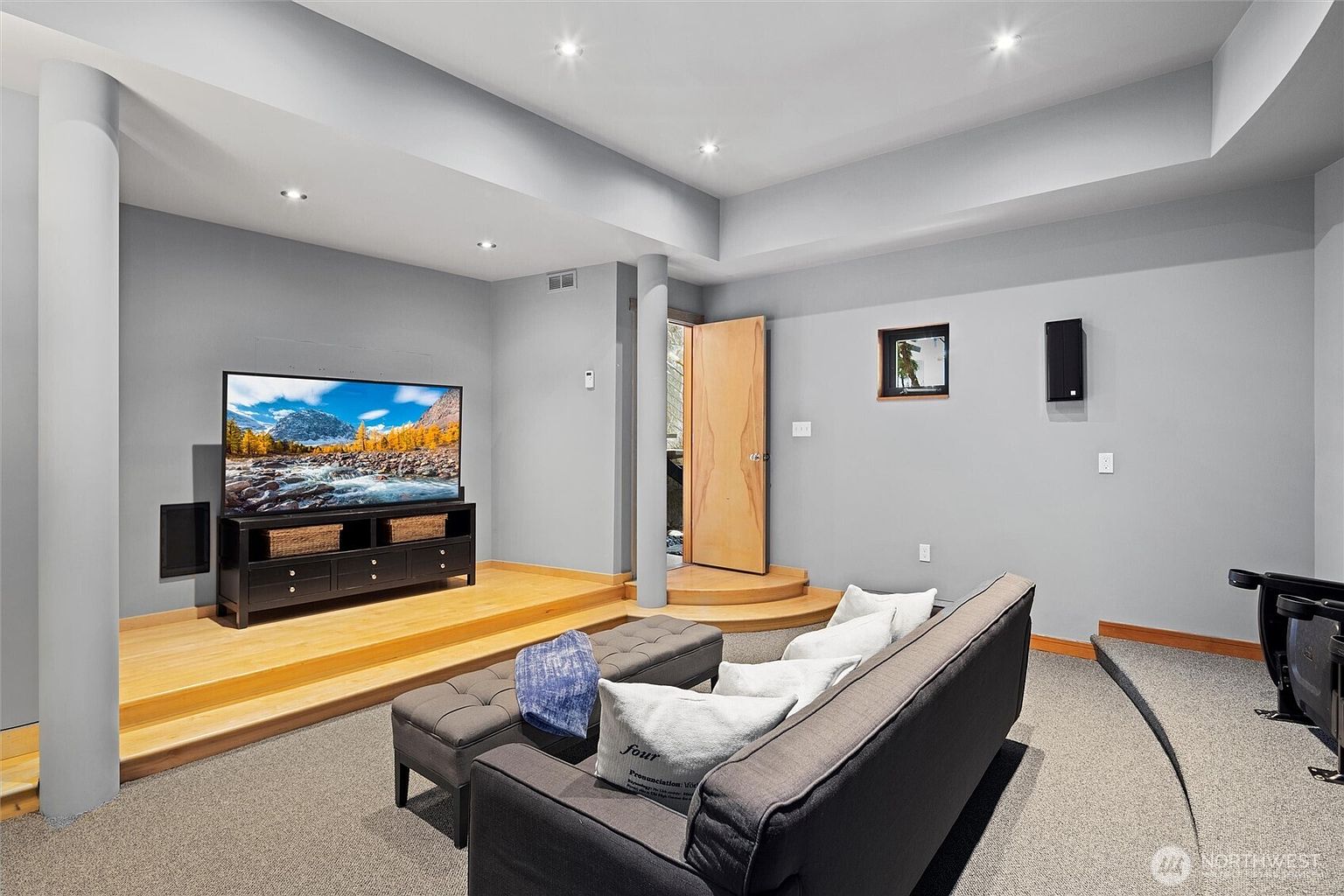 This spacious basement media room features a tiered layout with a raised wooden platform for the television and media console, creating a dedicated viewing area. The room is carpeted for comfort and includes a large, plush grey sofa with decorative pillows, perfect for relaxing. A neutral grey color palette, recessed lighting, and a clean, modern aesthetic make this an inviting space for entertainment.
