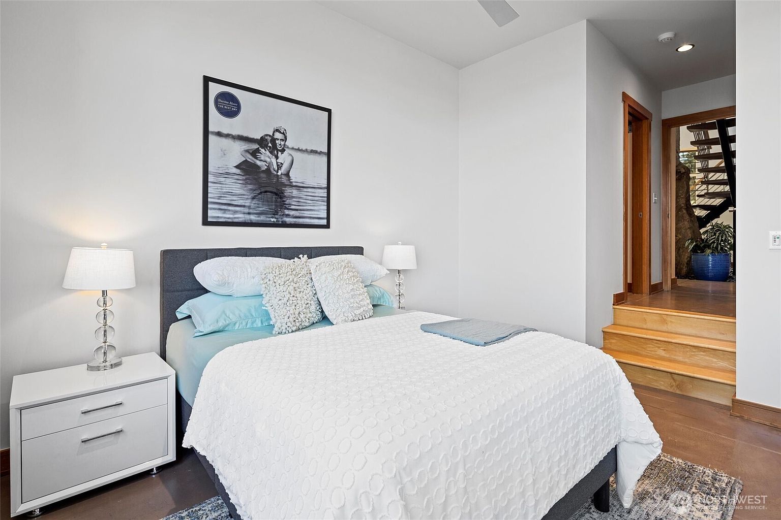 This cozy bedroom features a queen-sized bed with a grey upholstered headboard, dressed in light blue bedding and a textured white quilt. A white nightstand with a matching lamp sits to the left, while a framed black-and-white photograph hangs above the bed. The room leads to a small set of wooden stairs that open up to a view of a staircase and a potted plant, creating a unique architectural transition.