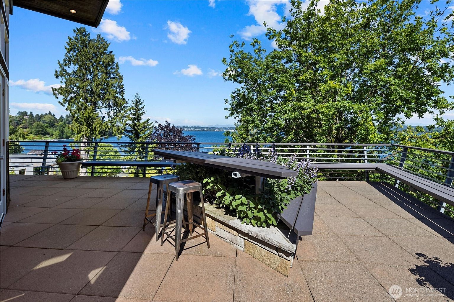 This expansive, elevated deck offers a stunning panoramic view of a serene lake surrounded by lush greenery and distant hills. The space features a built-in stone-based bench with a sleek, modern bar-style table, complemented by two industrial-style metal stools. The paved surface and clean lines create a sophisticated outdoor living area perfect for entertaining while enjoying the tranquil waterfront scenery.