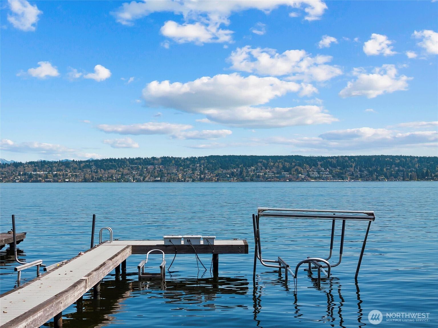This image captures a serene waterfront view from a private dock extending into a calm, expansive lake. In the foreground, a metal boat lift stands partially submerged in the water, while the background features a distant shoreline lined with trees and residential homes under a bright, cloud-filled sky. The perspective is a low-angle, eye-level shot from the end of the dock, emphasizing the peaceful and recreational nature of the property.