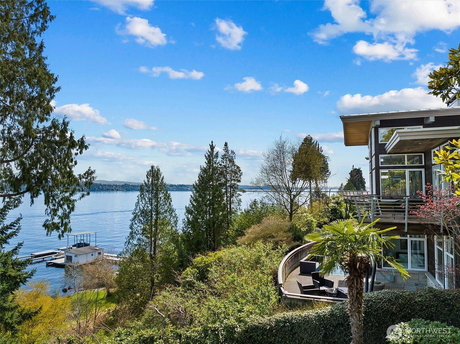 This stunning rear view of a modern, multi-level home showcases its seamless integration with the natural landscape and expansive waterfront views. The property features a tiered design with a private balcony, a curved patio area with outdoor seating, and lush greenery leading down to a private dock on the water. The architectural style emphasizes large windows and clean lines, creating a sophisticated and serene atmosphere perfect for luxury living.