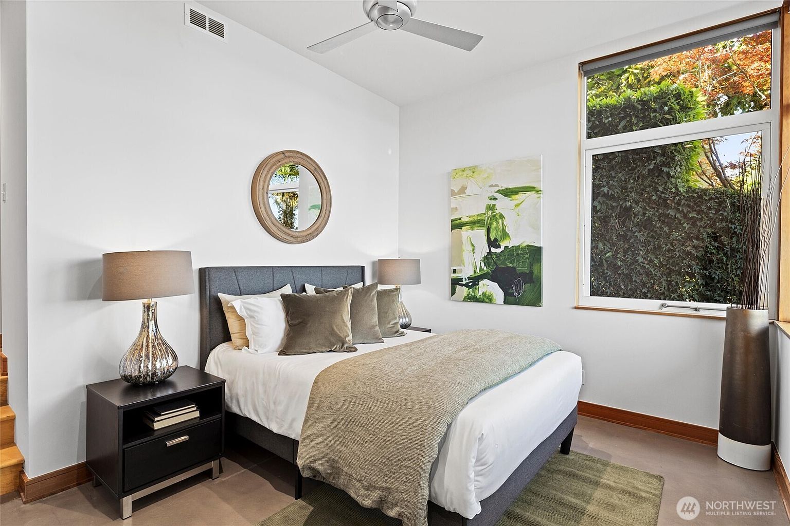This modern guest bedroom features a neutral color palette with a dark upholstered bed frame and a matching nightstand. A large window provides ample natural light and a view of lush greenery, while a contemporary abstract painting adds a pop of color to the wall. The room is completed by a ceiling fan and a circular mirror, creating a clean and inviting atmosphere.
