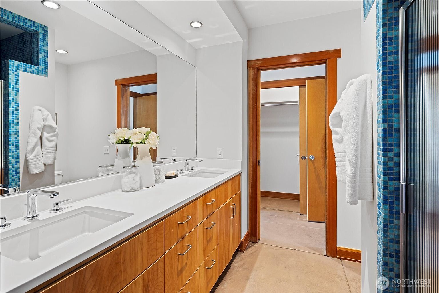 This primary bathroom features a long, warm-toned wood vanity with a clean white countertop and dual sinks, set against a large, frameless mirror. The space is accented by striking blue mosaic tile work around the shower and mirror frame, creating a vibrant contrast. A doorway leads directly into a walk-in closet, emphasizing a functional and well-connected layout.