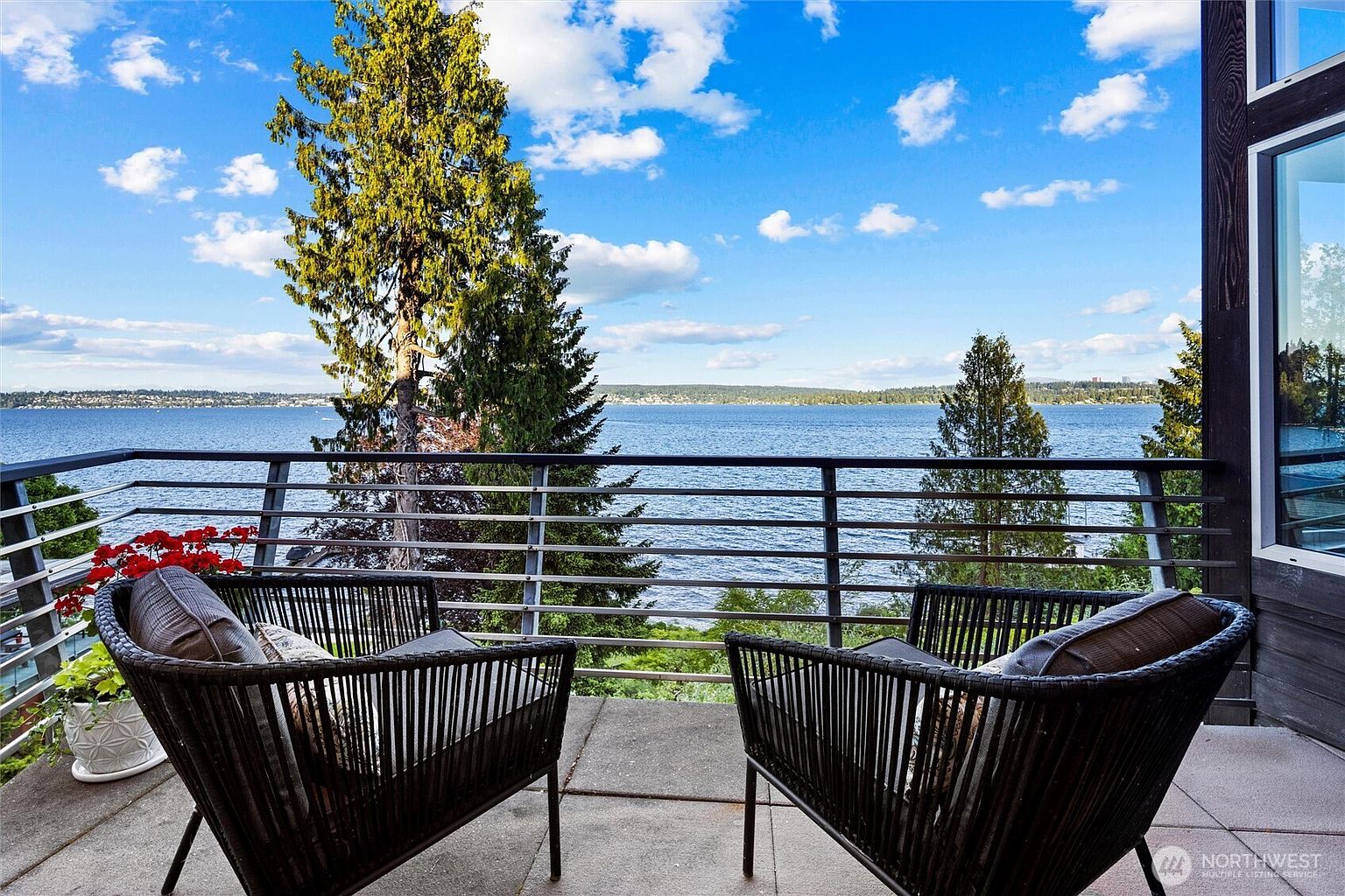 This elevated balcony offers a stunning, unobstructed panoramic view of a large lake under a bright, partly cloudy sky. The space is furnished with two modern, dark-wicker armchairs and features a sleek metal railing, creating a sophisticated and relaxing outdoor retreat. The perspective is from a seated position, emphasizing the serene connection between the private living space and the expansive natural scenery.