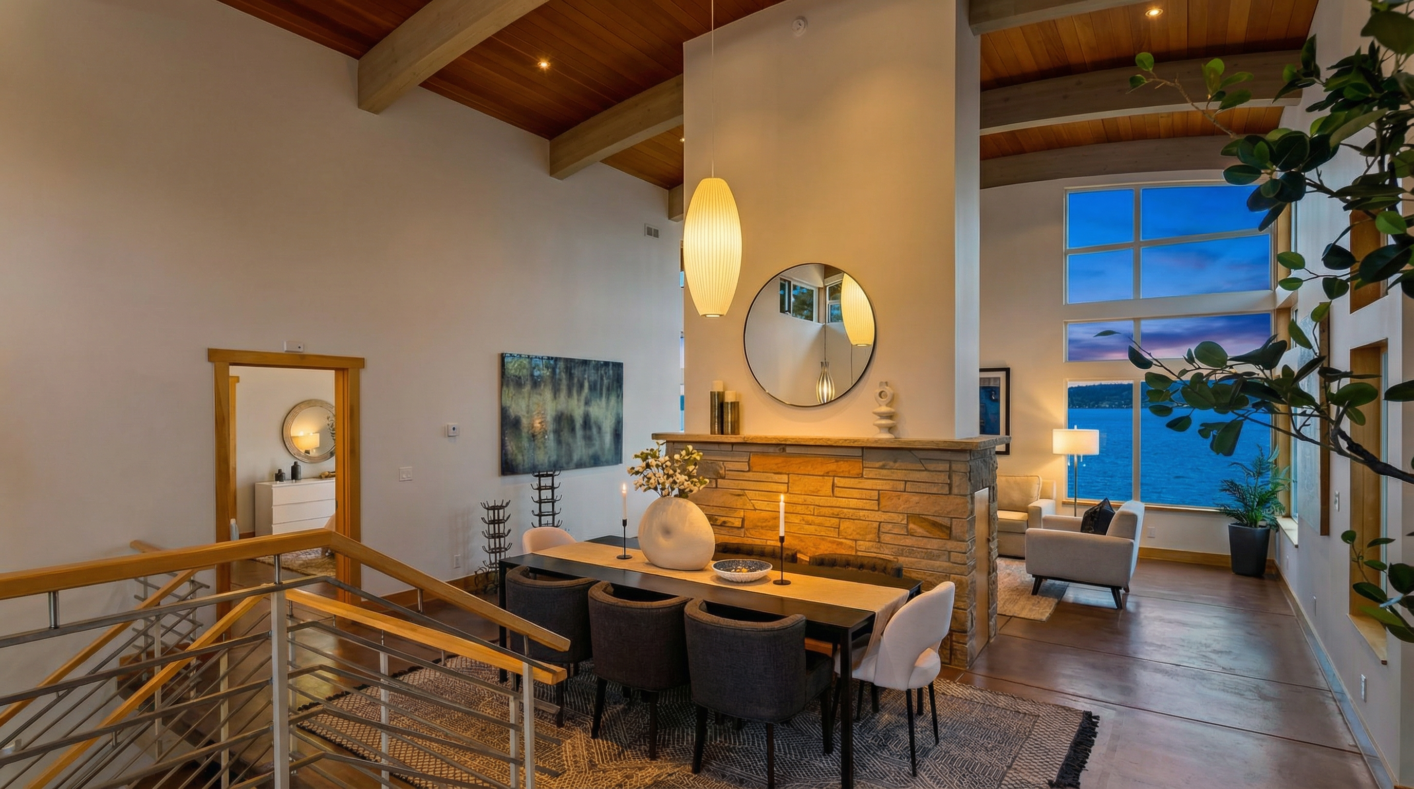 This elegant dining area features a contemporary table with six upholstered chairs, set beneath a striking modern pendant light and a high, vaulted wood-paneled ceiling with exposed beams. A central stone fireplace acts as a room divider, separating the dining space from a sunlit living area that offers expansive, panoramic views of the water through floor-to-ceiling windows. The open-concept design, combined with a neutral color palette and sophisticated decor, creates an airy and luxurious atmosphere.