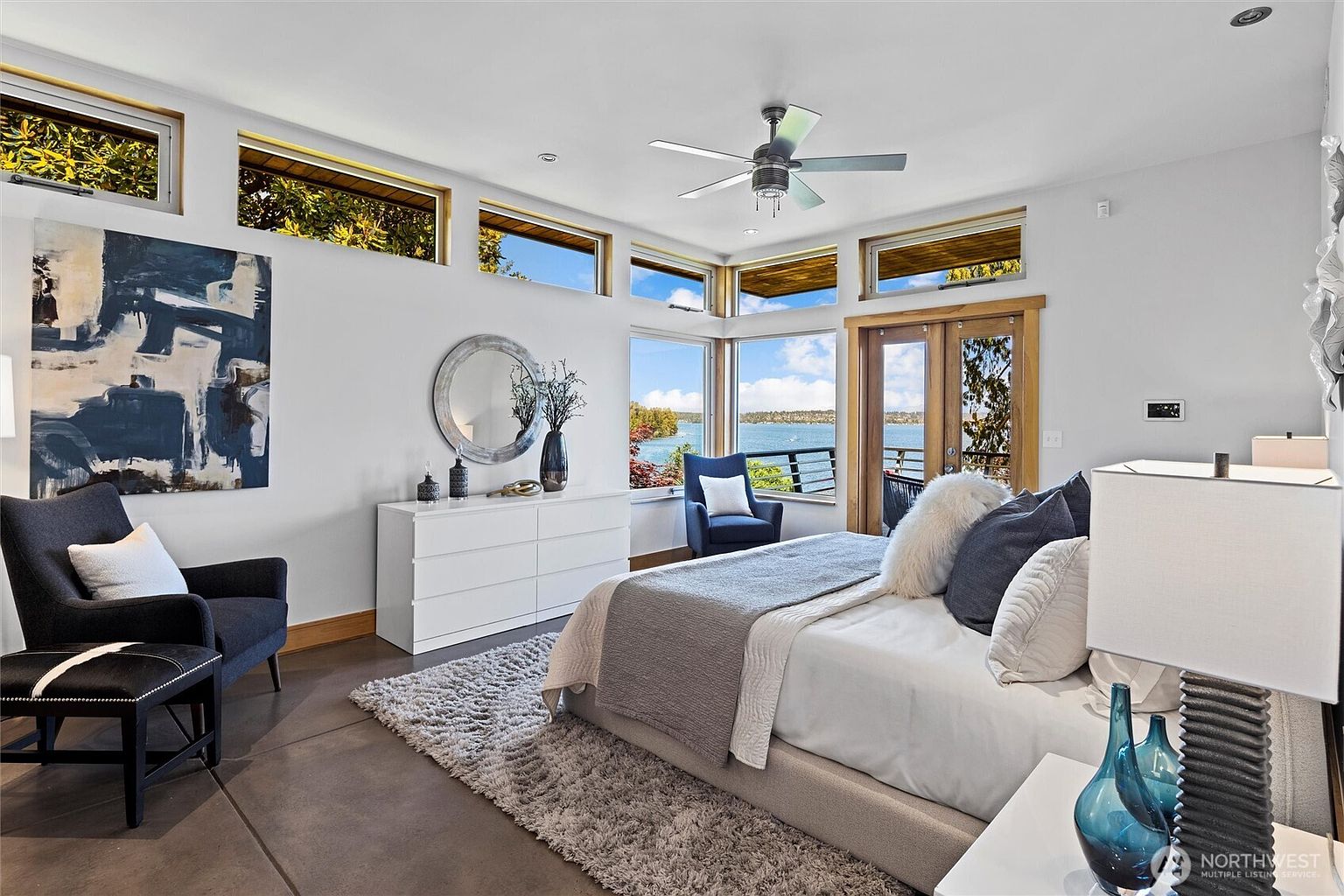 This bright and airy primary bedroom features a contemporary design with a neutral color palette, highlighted by a plush area rug and a modern bed frame. The room is framed by expansive windows that offer a stunning, panoramic view of the lake, seamlessly blending indoor comfort with the natural beauty outside. A sleek white dresser, a stylish armchair, and a ceiling fan complete the sophisticated, tranquil atmosphere of this waterfront retreat.