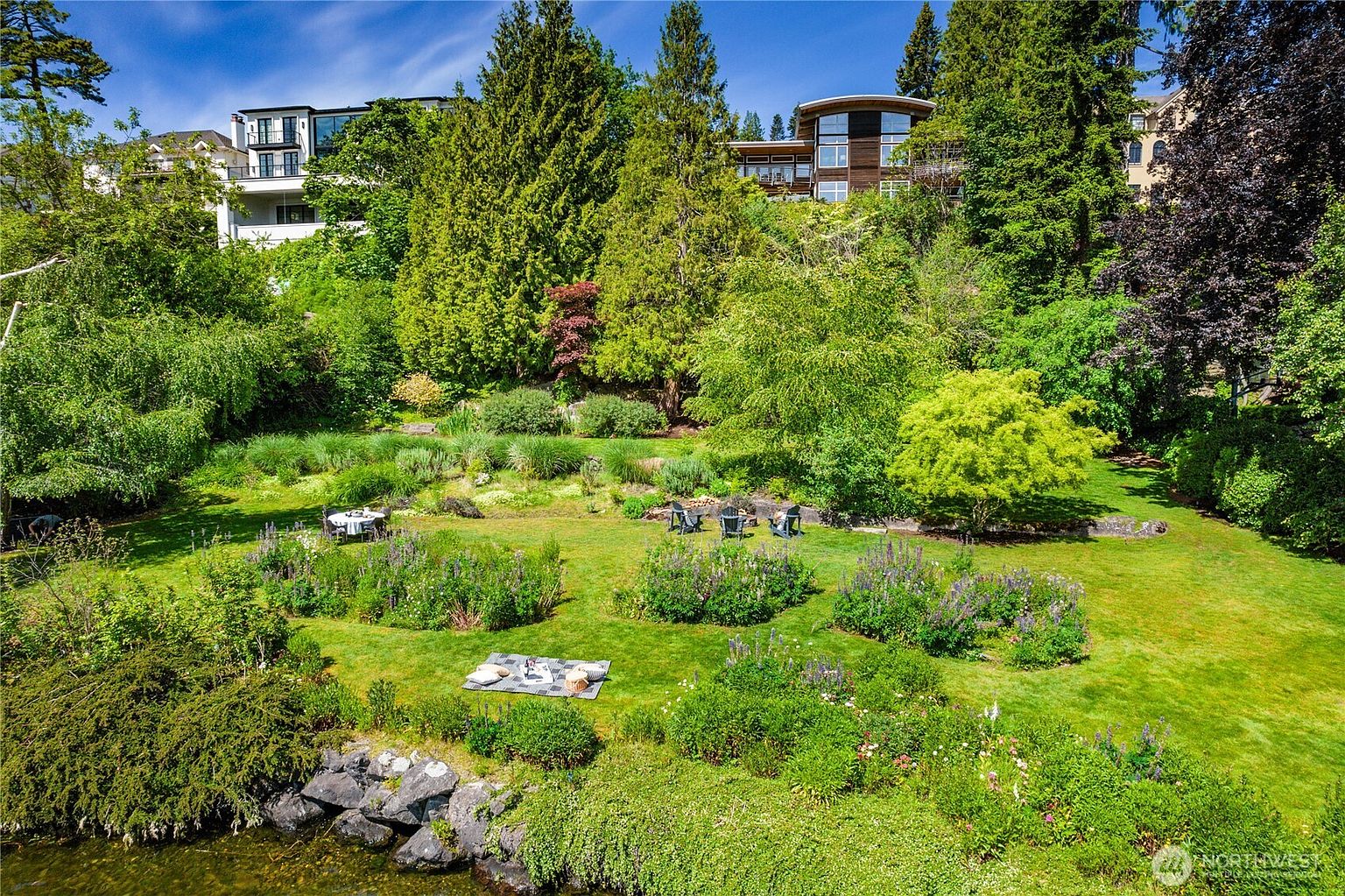 This expansive, lush backyard features a tiered landscape design with vibrant green lawns, mature trees, and well-maintained garden beds. A picnic blanket is spread out on the grass near the water's edge, while a small dining set and outdoor chairs are positioned further up the slope, creating a serene and inviting atmosphere. The perspective is elevated, looking down from the water toward the multi-level homes nestled into the hillside above.