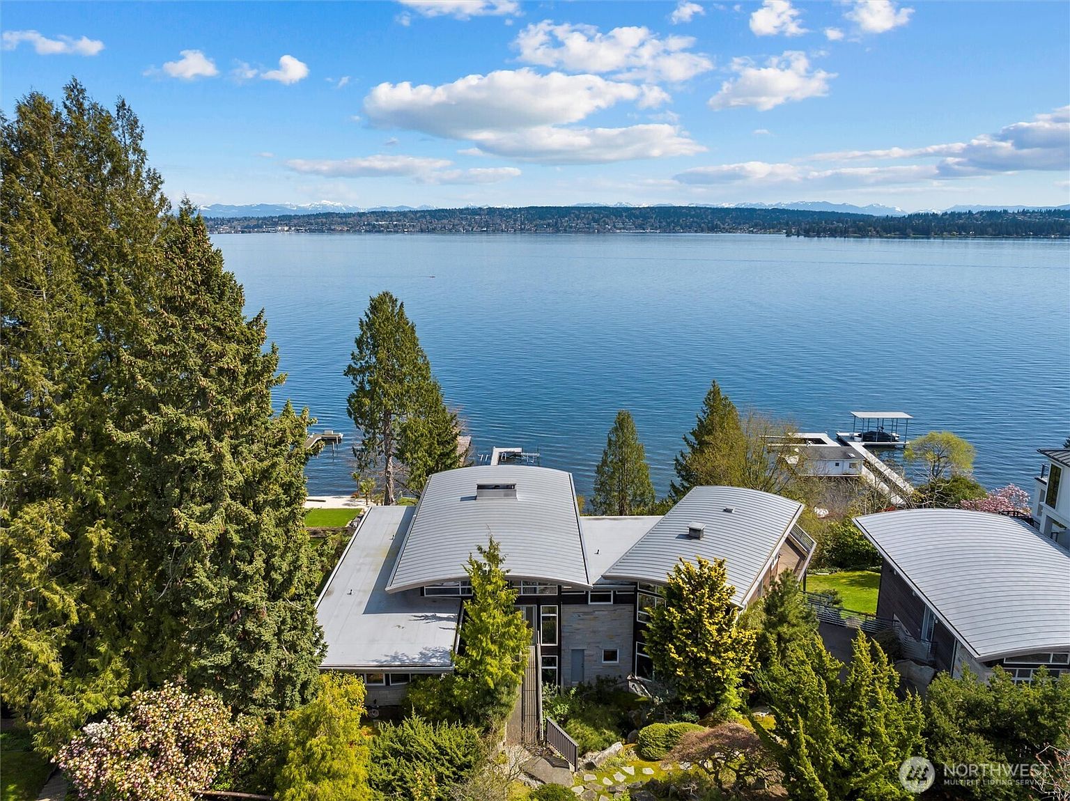 This high-angle aerial shot captures a contemporary waterfront residence featuring a distinctive, multi-sectioned curved metal roof. The home is nestled within a lush, mature landscape of evergreen trees, overlooking a serene, expansive lake with distant snow-capped mountains on the horizon. The perspective emphasizes the property's prime location, private dock, and seamless integration with the natural environment.