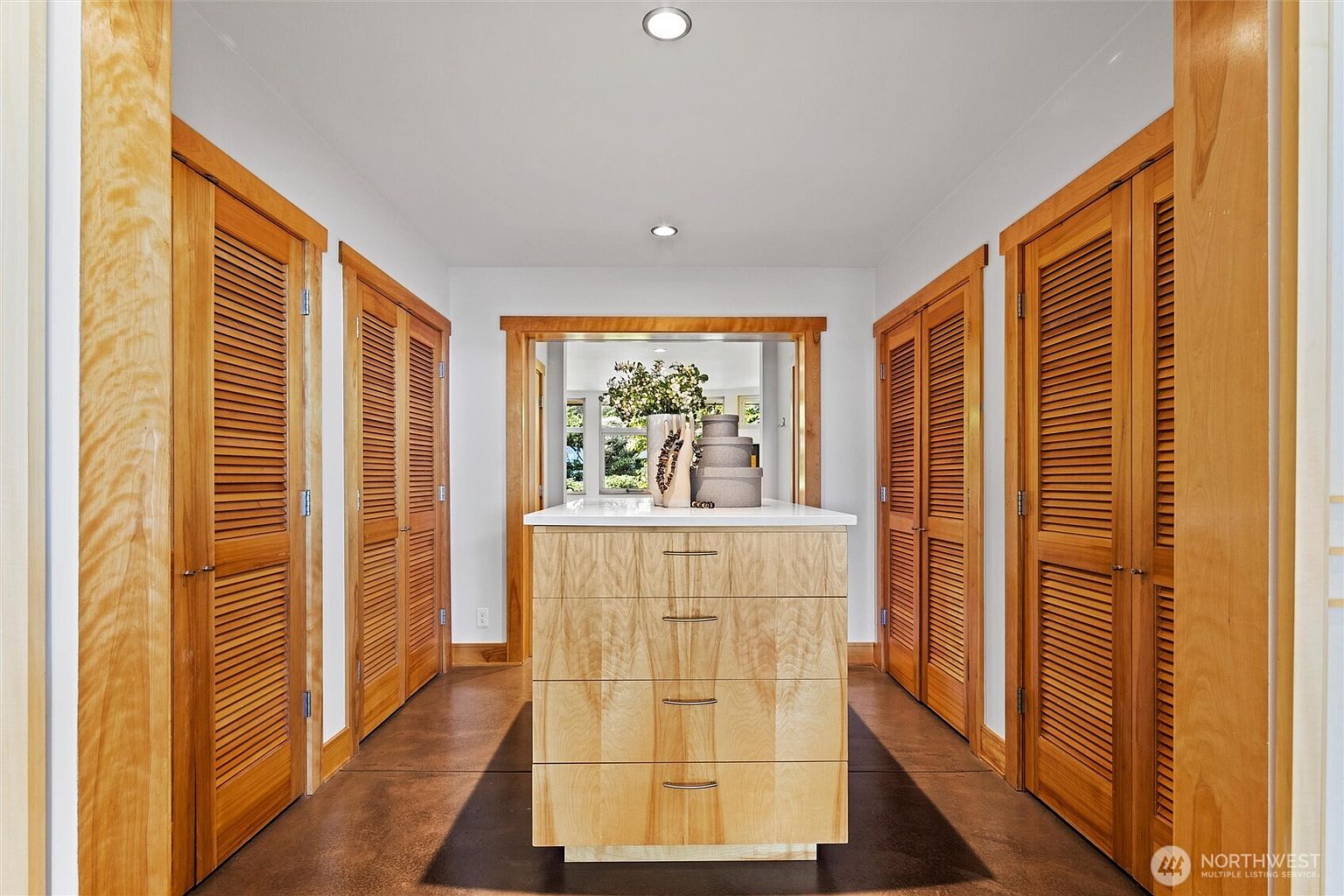 This spacious walk-in closet features a central island with light wood cabinetry and a white countertop, flanked by symmetrical rows of louvered wooden closet doors. The room is anchored by a dark, polished concrete floor that contrasts with the warm wood tones, while recessed ceiling lights provide a clean, modern illumination. The perspective is centered, drawing the eye through the closet toward an open doorway that reveals a bright, sunlit room beyond.