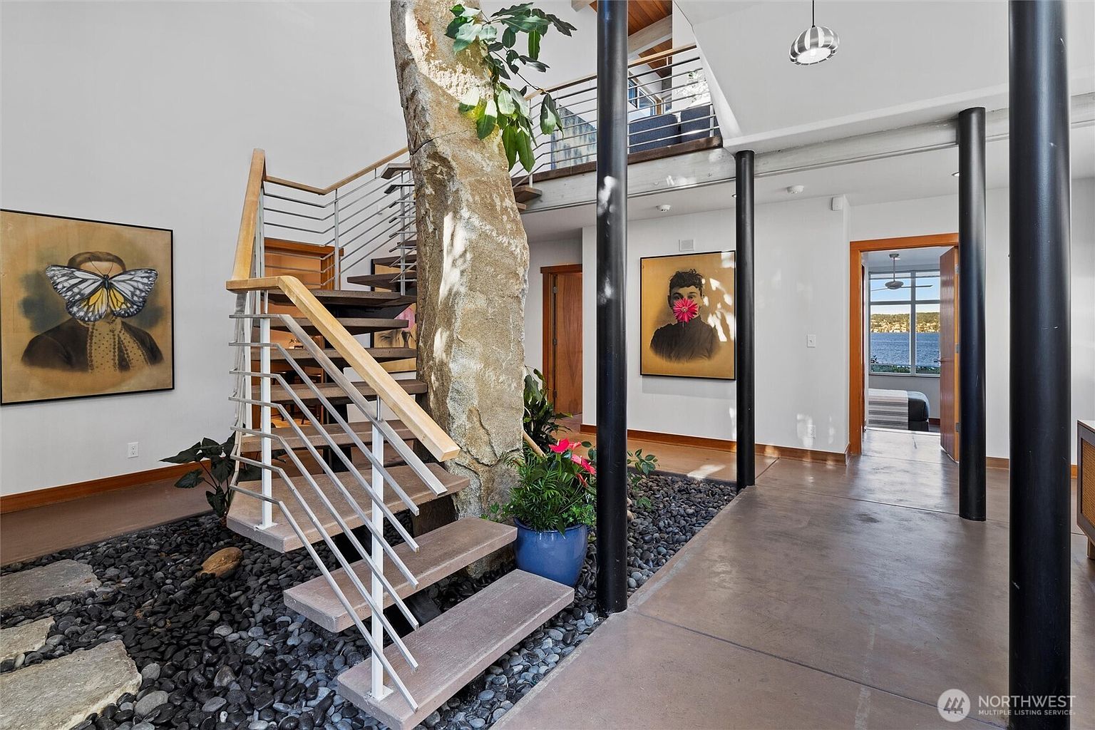 This unique interior space features a dramatic architectural centerpiece: a large, natural stone pillar integrated into the staircase design, surrounded by a decorative rock garden. The open-concept layout includes a modern floating staircase, sleek black support columns, and striking portrait artwork on the walls, creating a sophisticated, gallery-like atmosphere. The perspective looks through the foyer toward a bright doorway that reveals a scenic view of the water, emphasizing the home's connection to nature.
