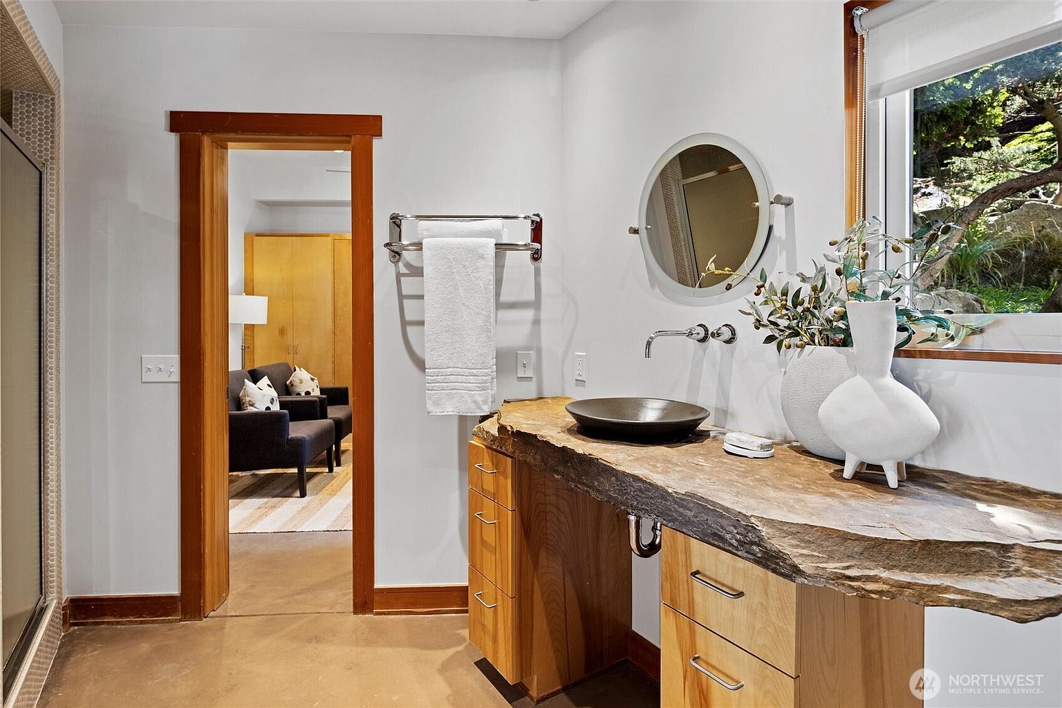 This bathroom features a striking live-edge wood vanity topped with a dark, bowl-style vessel sink and a wall-mounted faucet. A circular mirror hangs above the vanity, while a white towel hangs on a chrome rack nearby, and a view into an adjacent room reveals a cozy seating area. The space combines rustic natural textures with modern fixtures, creating a sophisticated and serene atmosphere.