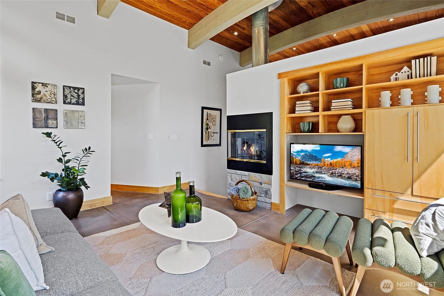 This inviting living room features a warm, contemporary aesthetic with a striking wood-paneled vaulted ceiling and exposed beams. The space is anchored by a cozy fireplace with a stone base, built-in light wood shelving, and a modern oval coffee table, all set against a neutral color palette. The perspective captures a comfortable seating area, highlighting the blend of natural textures and modern design elements that create a relaxed, upscale atmosphere.