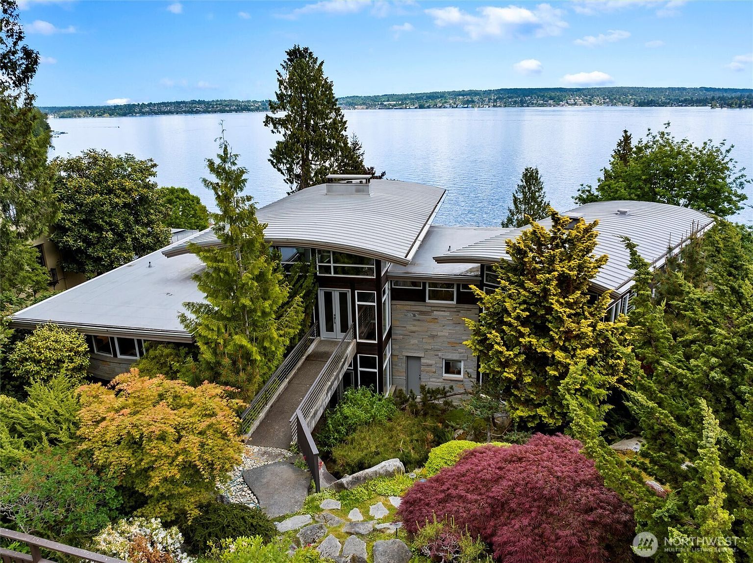 This striking contemporary home features a unique curved metal roof and a blend of stone and dark wood siding, nestled into a lush, tree-filled landscape overlooking a expansive lake. A pedestrian bridge leads directly to the elevated entryway, emphasizing the home's integration with the sloped terrain. The elevated perspective captures the architectural harmony between the modern structure and the serene waterfront setting.