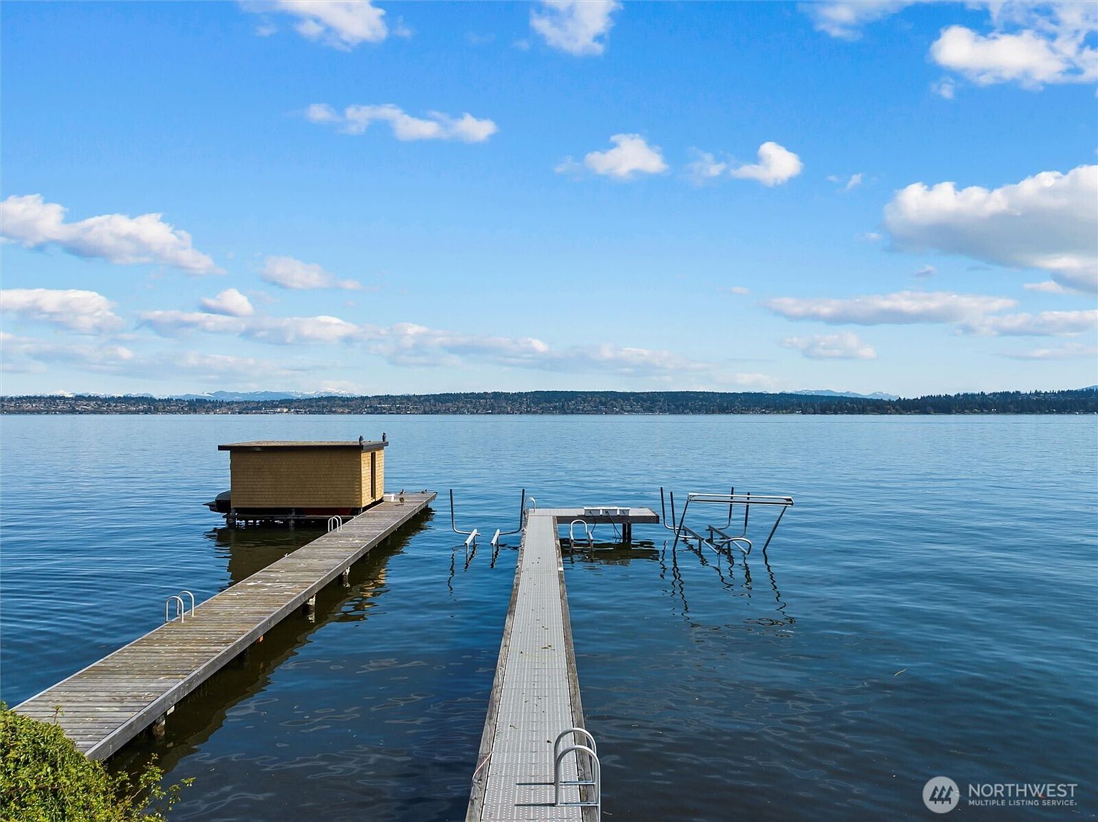 This scenic waterfront view features two parallel docks extending into a calm, expansive lake under a bright blue sky with scattered clouds. The left dock includes a small, enclosed wooden structure, while the right dock features metal boat lift frames, highlighting the property's direct water access. The perspective is a wide, eye-level shot that emphasizes the peaceful, recreational lifestyle offered by the waterfront location.