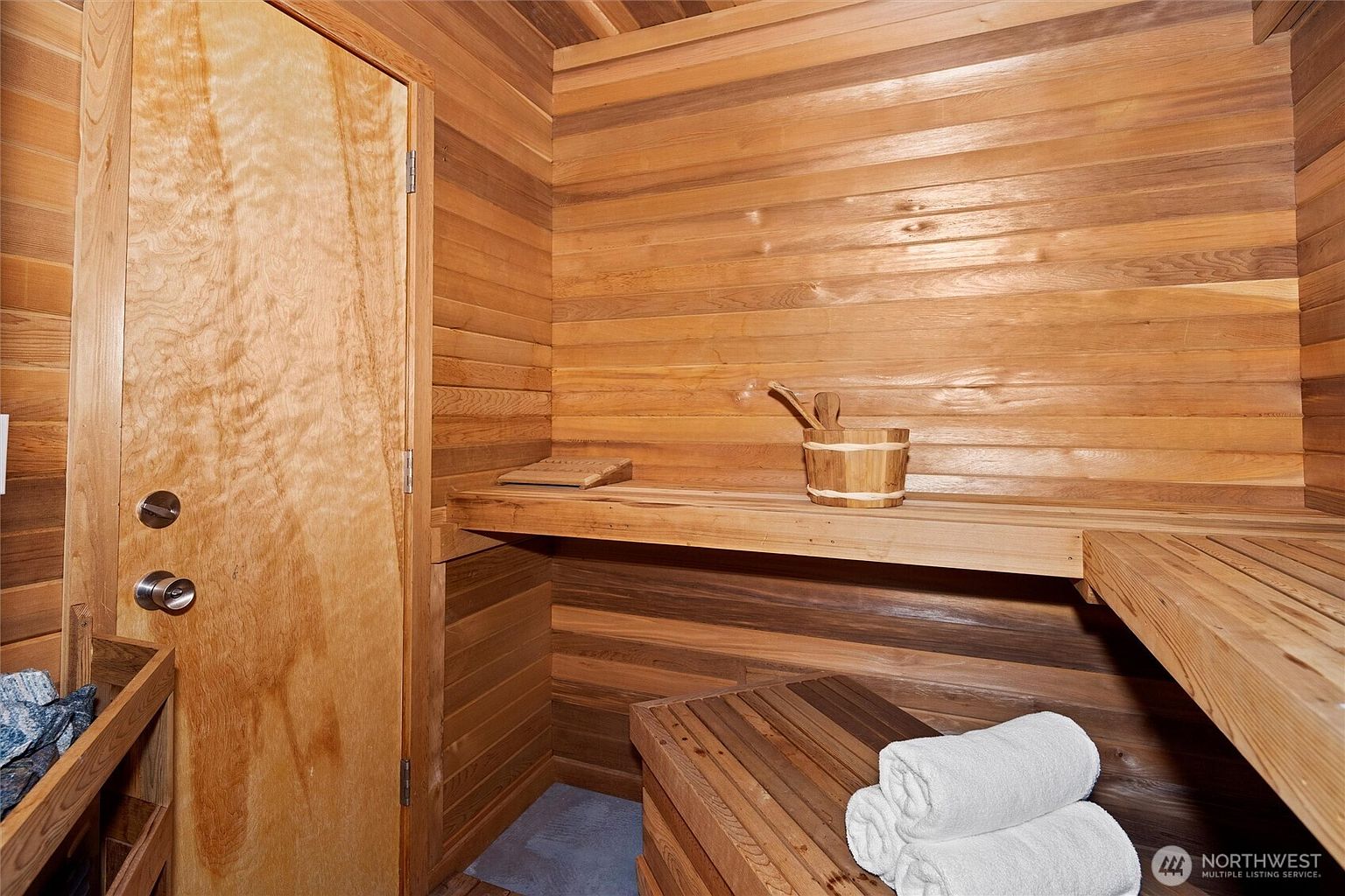 This image showcases a traditional cedar-lined sauna, featuring warm wood paneling throughout the walls and benches. A wooden bucket and ladle sit on the upper bench, while a stack of clean white towels rests on the lower bench, creating a relaxing and spa-like atmosphere. The perspective is from inside the sauna, looking toward the wooden door and the tiered seating arrangement.