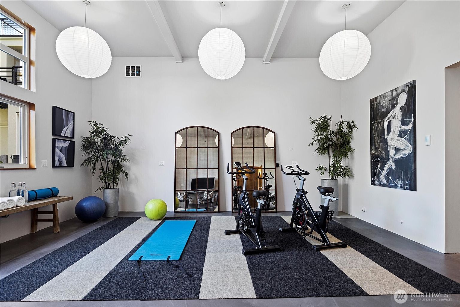 This spacious and modern home gym features high ceilings with three large, spherical pendant lights that create a bright and airy atmosphere. The room is equipped with two stationary exercise bikes, a yoga mat, a stability ball, and two large arched mirrors that enhance the sense of depth. A striking piece of abstract wall art and potted plants add a sophisticated, personal touch to this functional fitness space.