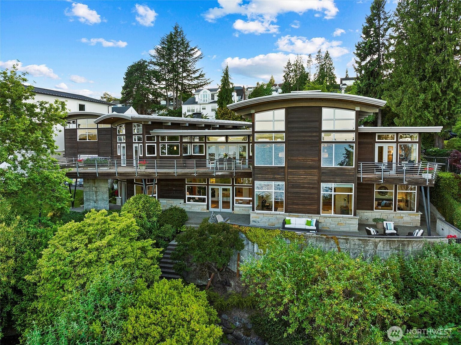 This stunning contemporary residence features a multi-level design with dark wood siding, expansive floor-to-ceiling windows, and a prominent curved roofline. The home includes a spacious elevated deck and a lower-level stone patio, all set against a lush, wooded landscape that suggests a serene, private setting. The perspective is a slightly elevated eye-level shot, capturing the architectural harmony between the modern structure and the natural environment.