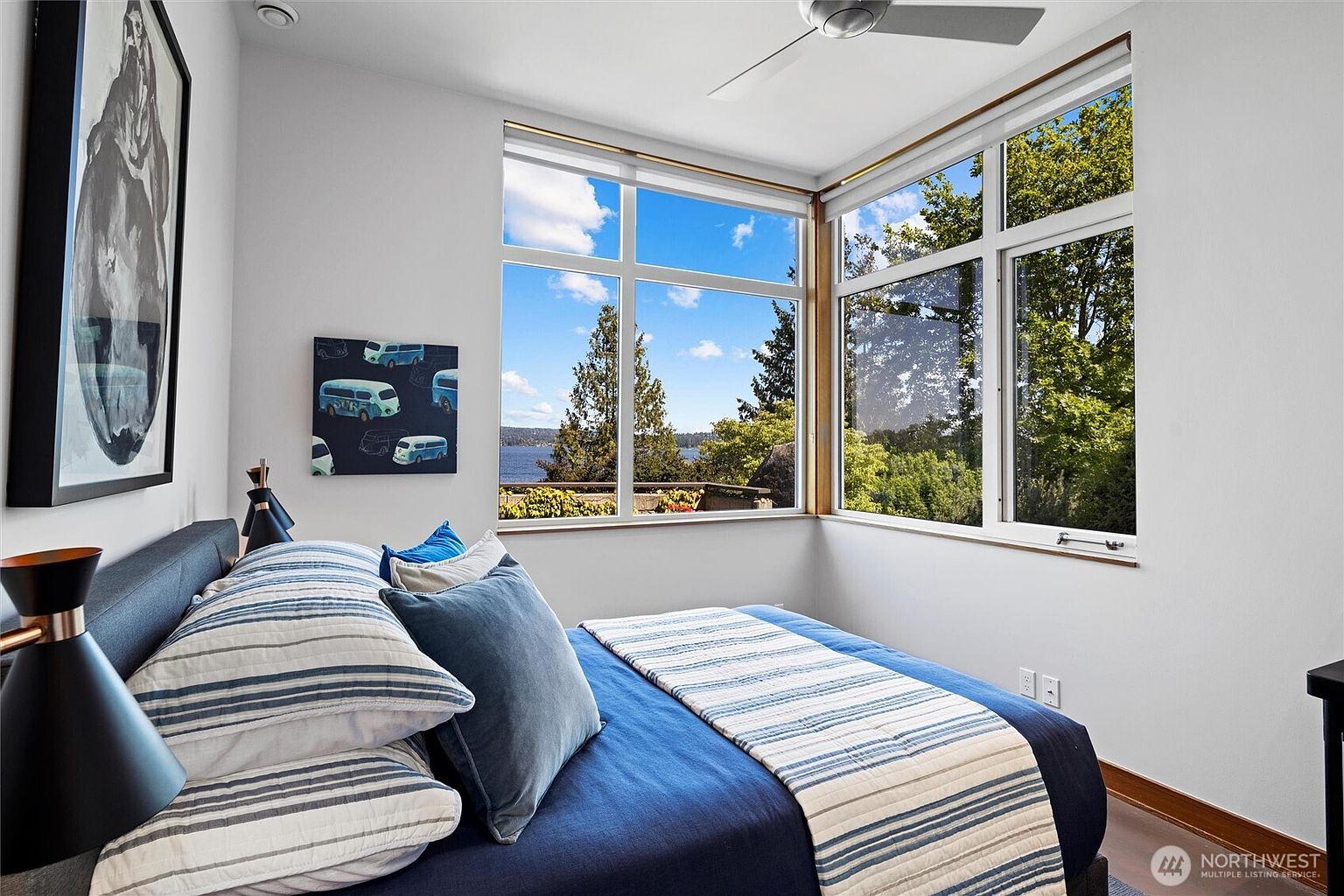 This modern bedroom features a cozy bed dressed in blue and white striped bedding, positioned against a wall adorned with contemporary artwork. Large corner windows flood the space with natural light and offer a serene, elevated view of the surrounding trees and water. The room's clean lines and minimalist aesthetic create a tranquil and inviting atmosphere.