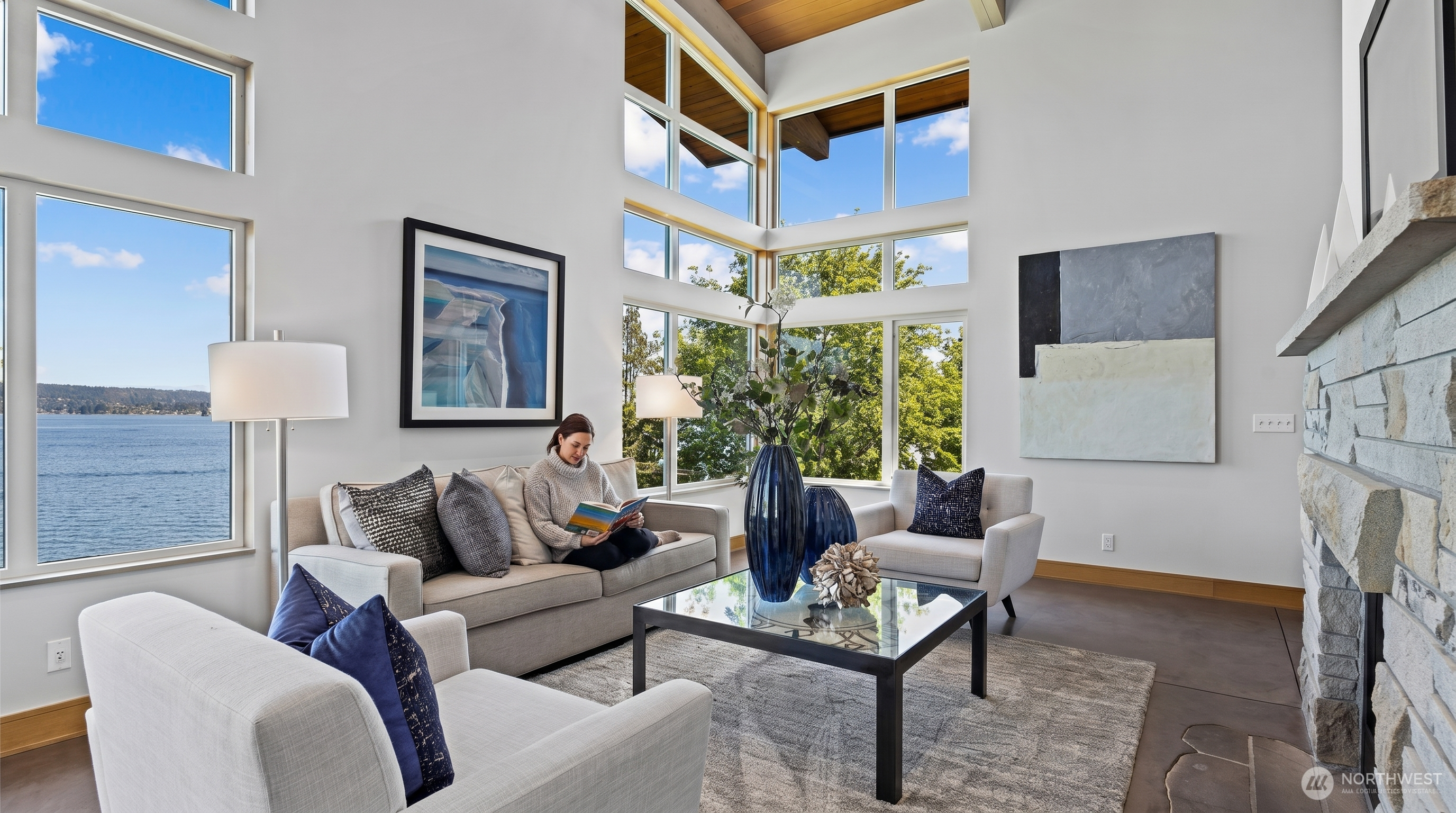 This bright and airy living room features soaring vaulted ceilings with wood paneling and expansive floor-to-ceiling windows that offer a stunning, unobstructed view of the water. The space is furnished with a neutral-toned sofa and armchairs, accented by deep blue pillows and a glass-topped coffee table, all centered around a rustic stone fireplace. The overall aesthetic is a sophisticated blend of modern luxury and natural warmth, perfectly framing the scenic outdoor landscape.