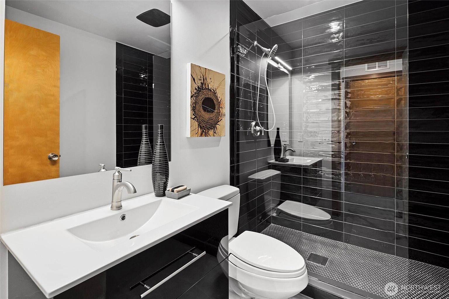 This modern bathroom features a sleek vanity with a white countertop, a black cabinet, and a contemporary faucet. The shower area is enclosed by a glass door and showcases striking black horizontal tiles contrasted with a textured pebble-style floor. A piece of abstract art hangs on the wall, adding a touch of personality to the minimalist, high-contrast design.