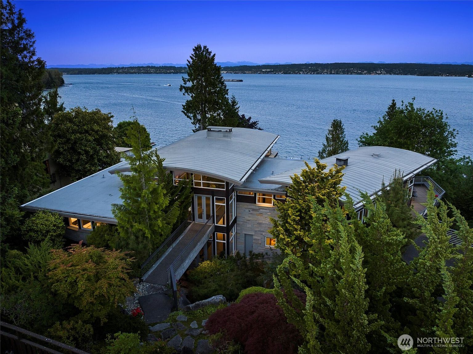 This stunning aerial perspective captures a contemporary waterfront residence featuring unique, curved metal roofing and a blend of stone and dark siding. A pedestrian bridge leads to the main entrance, nestled amidst lush, mature landscaping that slopes down toward the expansive water view. The twilight lighting enhances the architectural elegance of the home, creating a serene and sophisticated atmosphere.