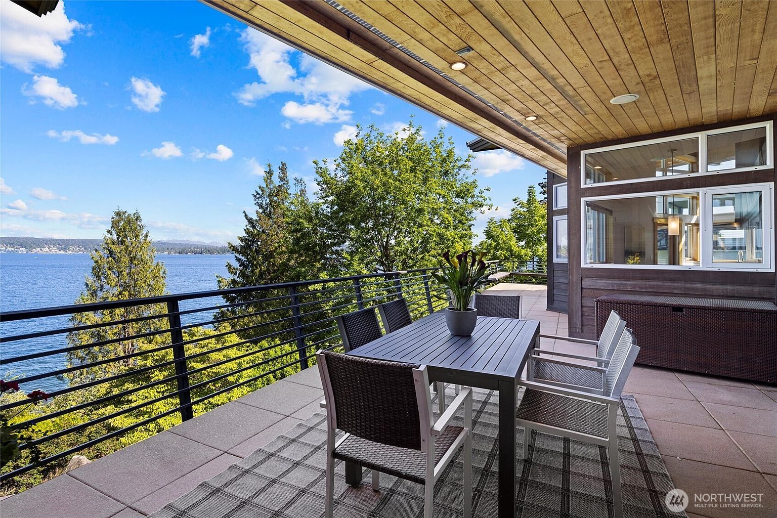 This expansive, covered deck offers a stunning, elevated view of a large lake, framed by lush green trees and a clear blue sky. The space is furnished with a modern dining set placed on a patterned outdoor rug, creating an inviting area for al fresco dining. The architectural design features warm wood-paneled ceilings with recessed lighting, seamlessly blending indoor comfort with the natural beauty of the waterfront setting.