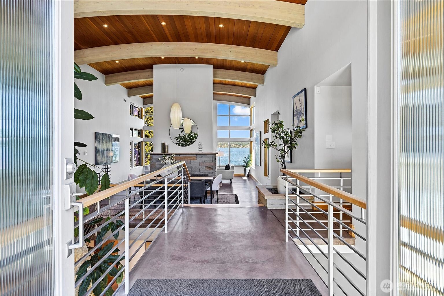 This striking entryway features a bridge-like walkway with modern metal railings that leads into an expansive, open-concept living space with vaulted wood-paneled ceilings. The perspective from the front door highlights a seamless transition into the home, showcasing a stone fireplace, contemporary dining area, and floor-to-ceiling windows that frame a stunning waterfront view. The design aesthetic is sophisticated and airy, emphasizing architectural depth and natural light.