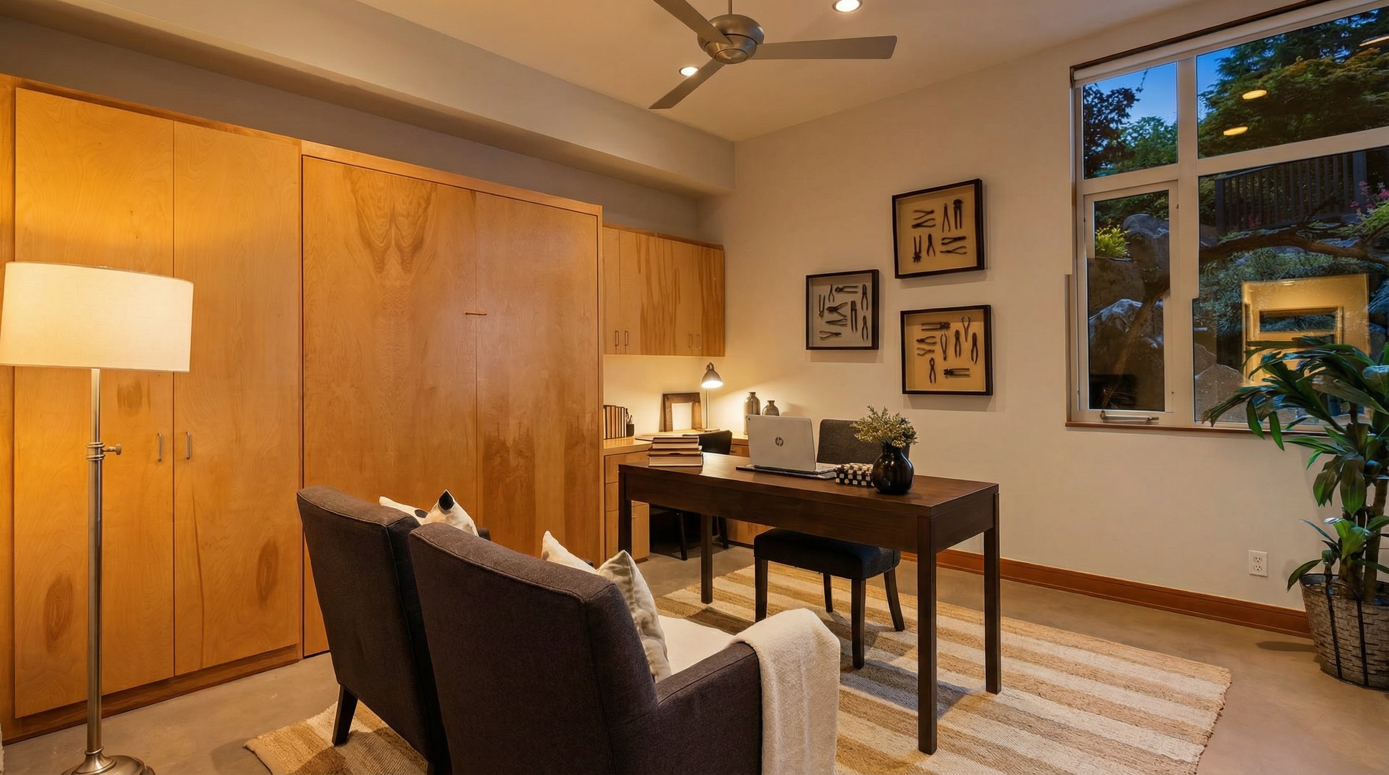 This well-appointed home office features a sleek, dark wood desk paired with a matching chair, set against a backdrop of light wood cabinetry and a Murphy bed unit. The room is accented by a striped area rug, a floor lamp, and framed tool-themed artwork, creating a professional yet inviting atmosphere. A large window provides natural light and a view of the outdoor landscape, enhancing the bright and airy feel of the workspace.