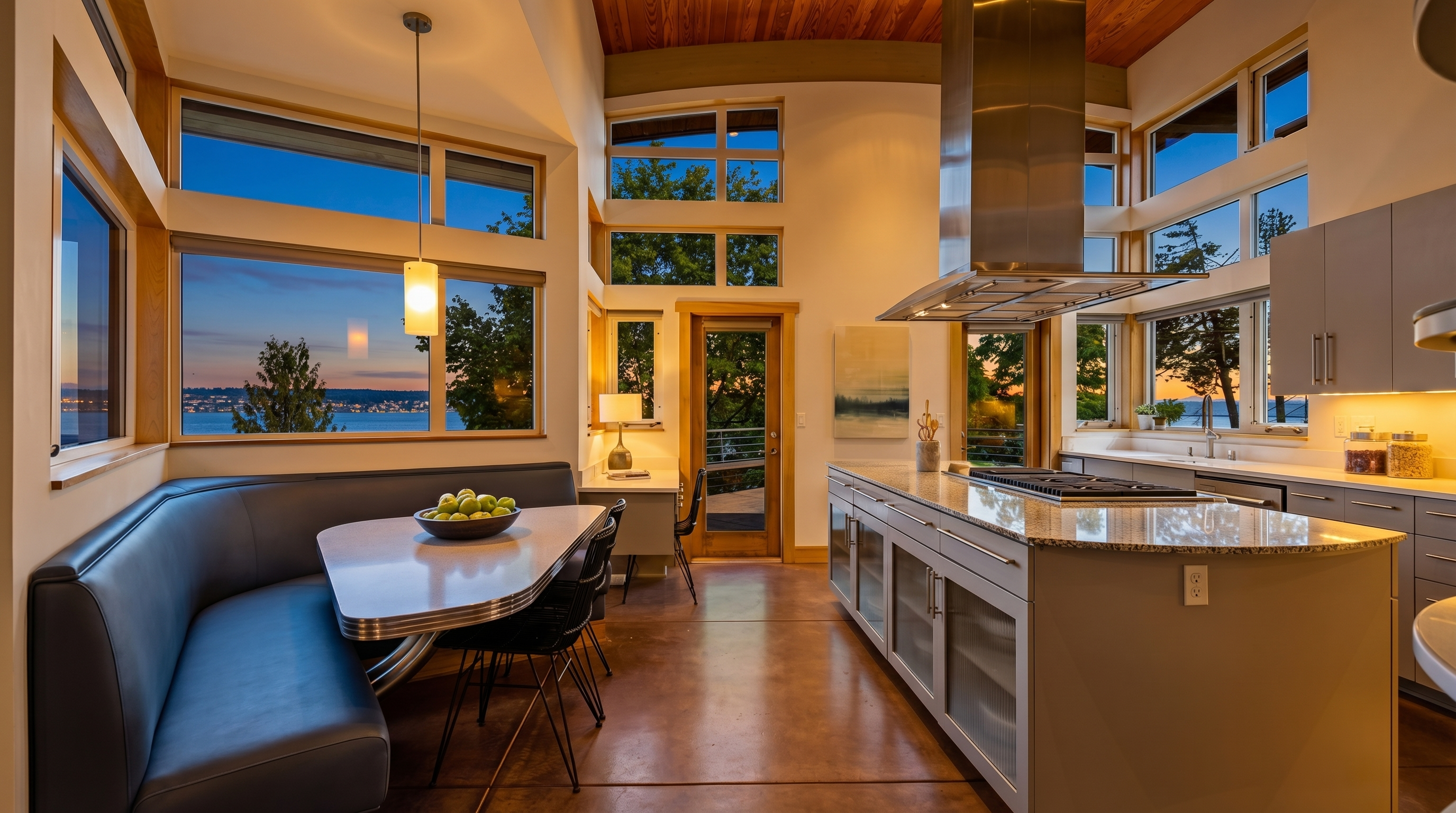 This modern, open-concept kitchen features a large central island with a gas cooktop and a sleek stainless steel range hood, complemented by a built-in dining nook with a curved blue banquette. The space is defined by high, wood-paneled ceilings, polished concrete floors, and expansive windows that offer panoramic water views and abundant natural light. The design seamlessly blends contemporary industrial elements with warm, natural textures, creating an airy and sophisticated atmosphere.