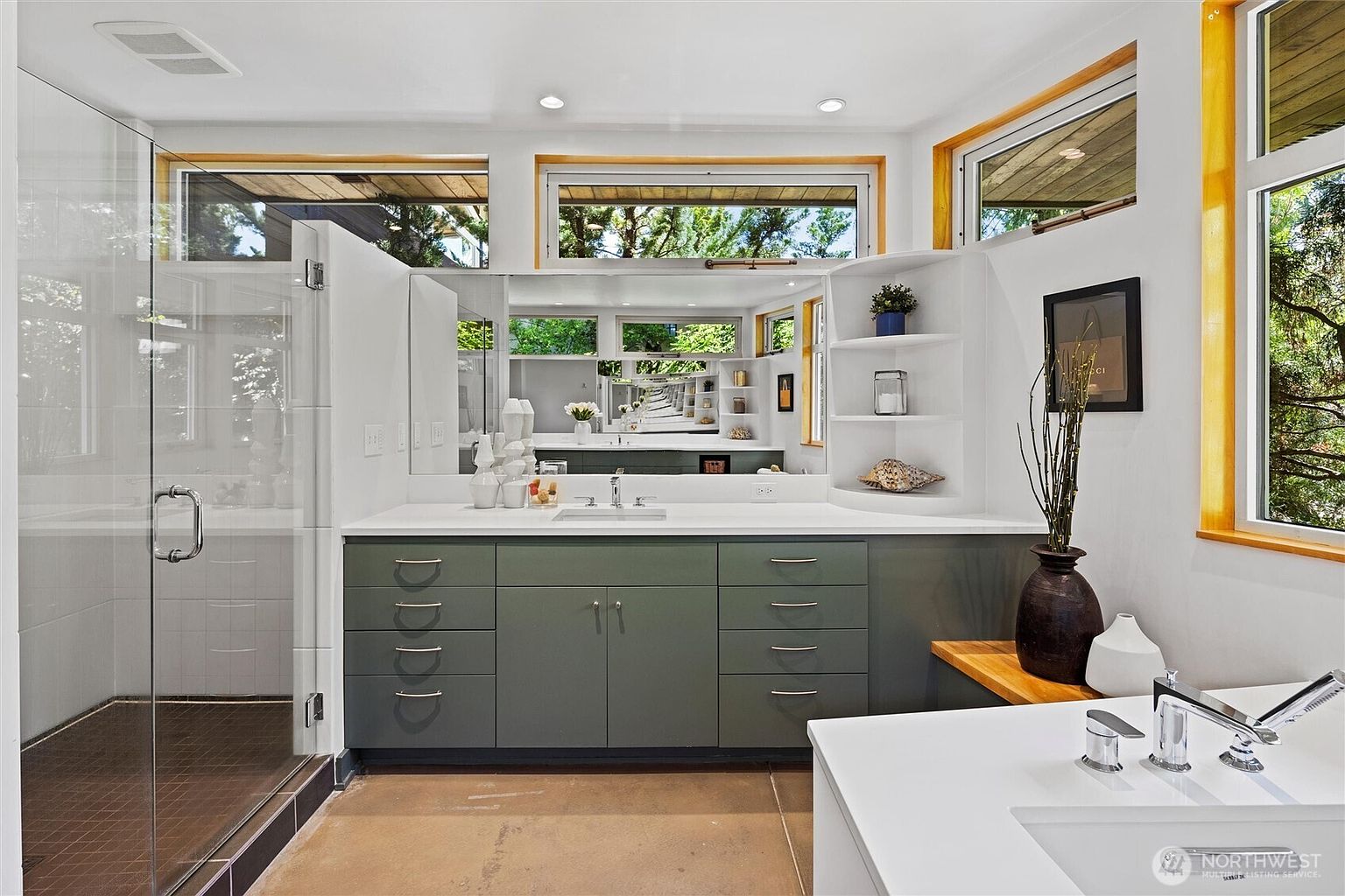 This modern primary bathroom features a sleek, sage-green vanity with a white countertop and integrated sink, complemented by a large mirror and built-in shelving. To the left, a spacious glass-enclosed walk-in shower adds a contemporary touch, while large windows allow for abundant natural light and views of the surrounding greenery. The overall aesthetic is clean, minimalist, and serene, emphasizing a bright and airy atmosphere.