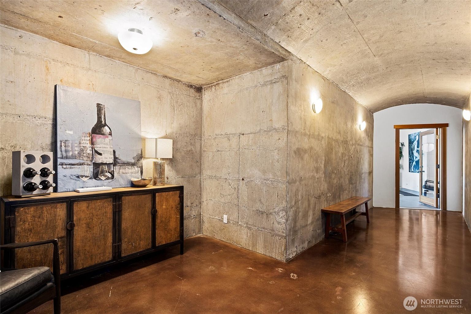 This unique basement space features raw, industrial-style concrete walls and a vaulted ceiling, creating a dramatic and moody atmosphere. The room is furnished with a rustic wooden sideboard topped with a wine-themed art piece and a lamp, while a simple wooden bench sits along the curved wall. The polished concrete floor reflects the warm glow of the wall-mounted sconces, leading the eye toward an open doorway that reveals a brighter, more traditional living area beyond.