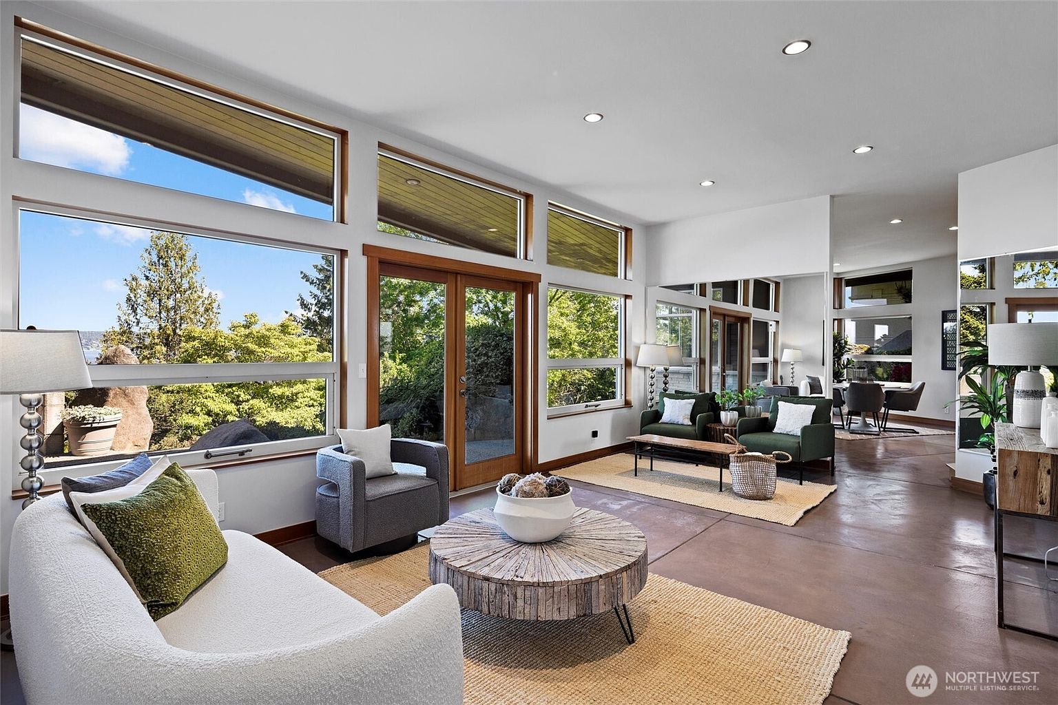 This bright and airy living room features a contemporary design with expansive floor-to-ceiling windows that offer a scenic view of the outdoors. The space is furnished with a textured white sofa, a rustic circular wooden coffee table, and two dark green armchairs, all set upon natural fiber rugs over a polished concrete floor. A large mirror on the wall enhances the sense of space, reflecting the adjacent dining area and creating a seamless, open-concept flow.