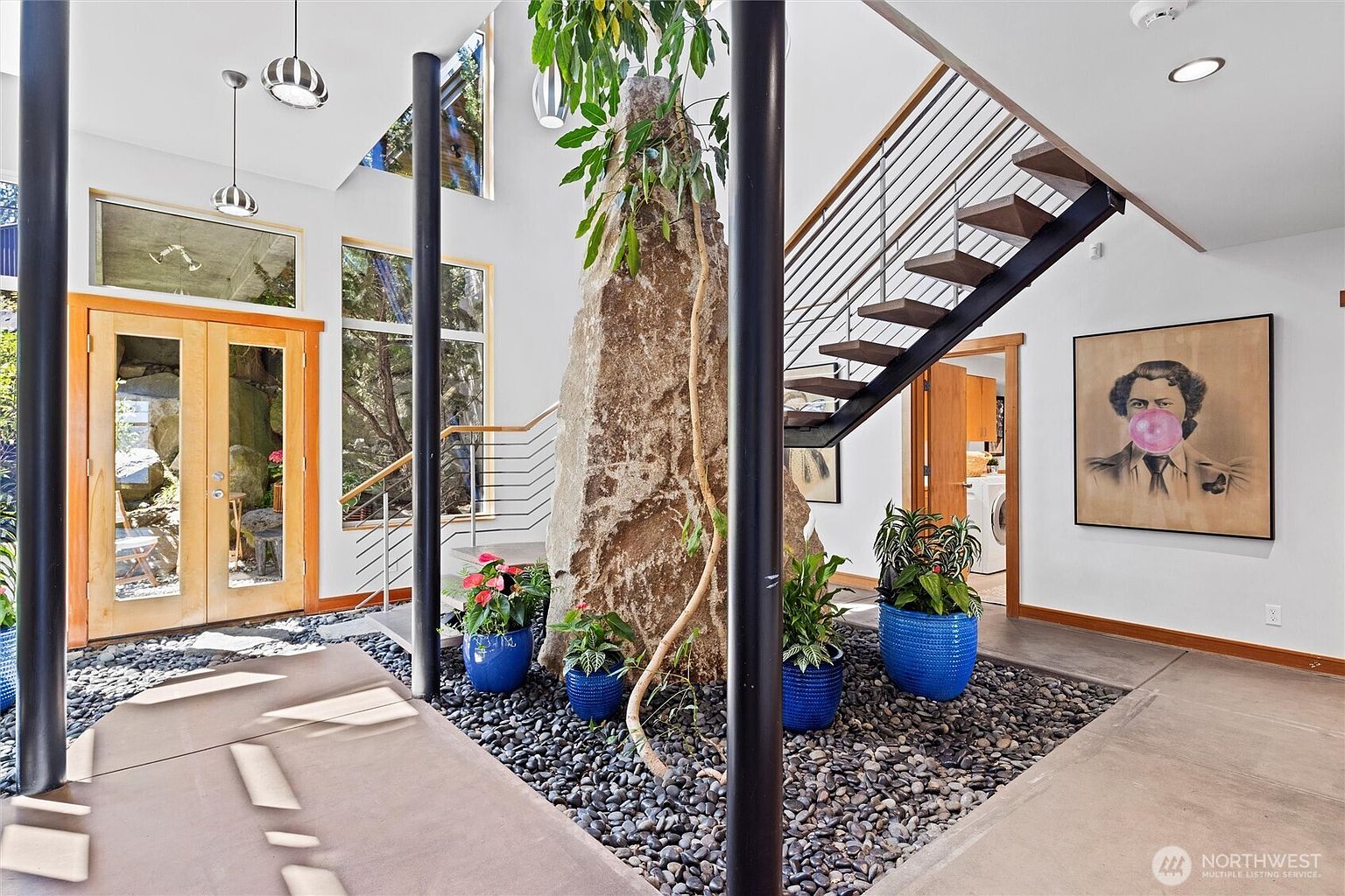 This striking entryway features a dramatic, double-height foyer centered around a massive, natural rock formation integrated into the home's design. A modern staircase with open treads and metal railings ascends alongside the rock, while a bed of dark river stones and vibrant blue planters filled with lush greenery create a serene, organic atmosphere. The space is illuminated by contemporary pendant lighting and natural light streaming through large windows, showcasing a unique blend of architectural boldness and natural elements.