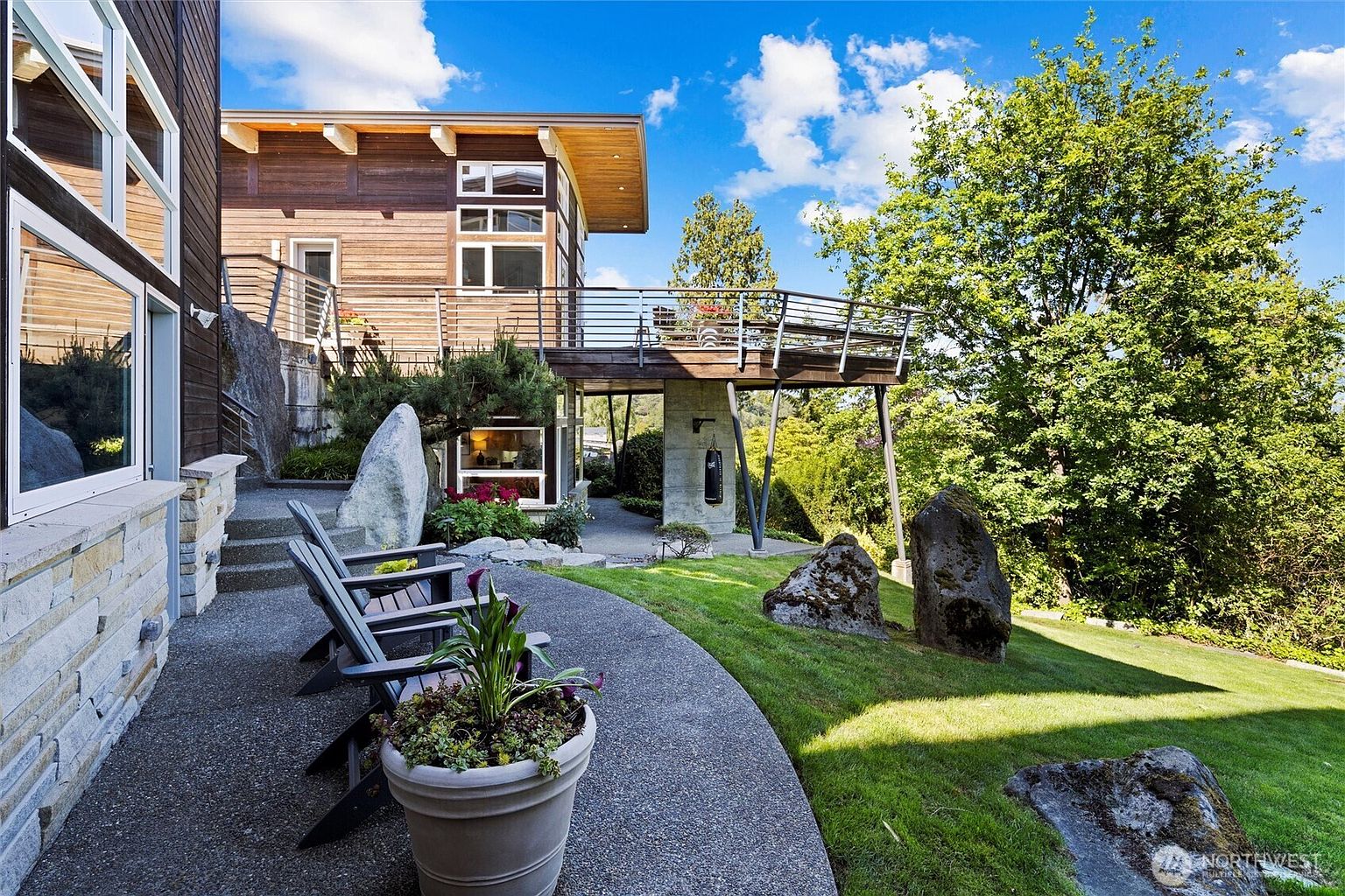This stunning contemporary home features a multi-level design with a prominent elevated deck supported by sleek metal pillars, overlooking a beautifully landscaped backyard. The exterior combines warm wood siding with stone accents, creating a sophisticated architectural aesthetic that blends seamlessly with the natural rocky terrain. A curved concrete patio with Adirondack chairs provides a perfect vantage point to enjoy the lush green lawn and mature trees.