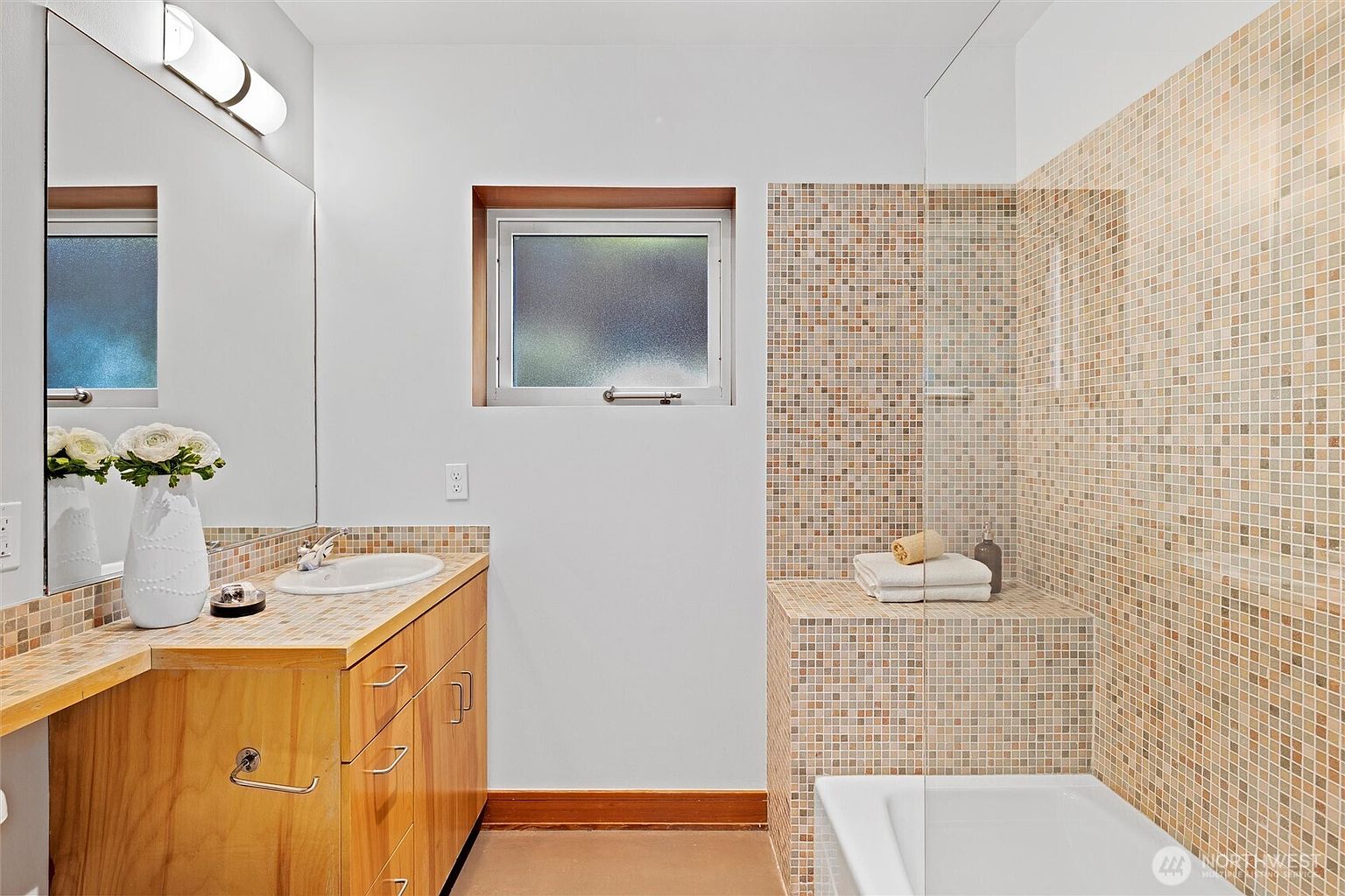 This bathroom features a warm, earthy aesthetic with mosaic tile accents on the vanity countertop and shower surround. The space includes a wooden vanity, a large wall-mounted mirror, and a glass-enclosed shower area with a built-in tiled bench. A frosted window provides natural light while maintaining privacy, creating a functional and cozy atmosphere.