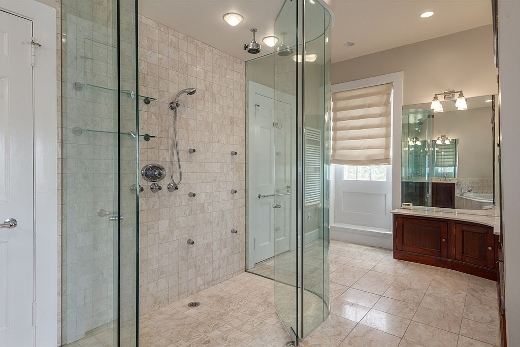 This primary bathroom features a spacious walk-in shower with a curved glass enclosure and light-toned stone tile walls. A dark wood vanity with a stone countertop and dual mirrors sits to the right, complemented by warm lighting fixtures. The room is finished with light-colored floor tiles and a window covered by a neutral Roman shade, creating a bright and functional space.