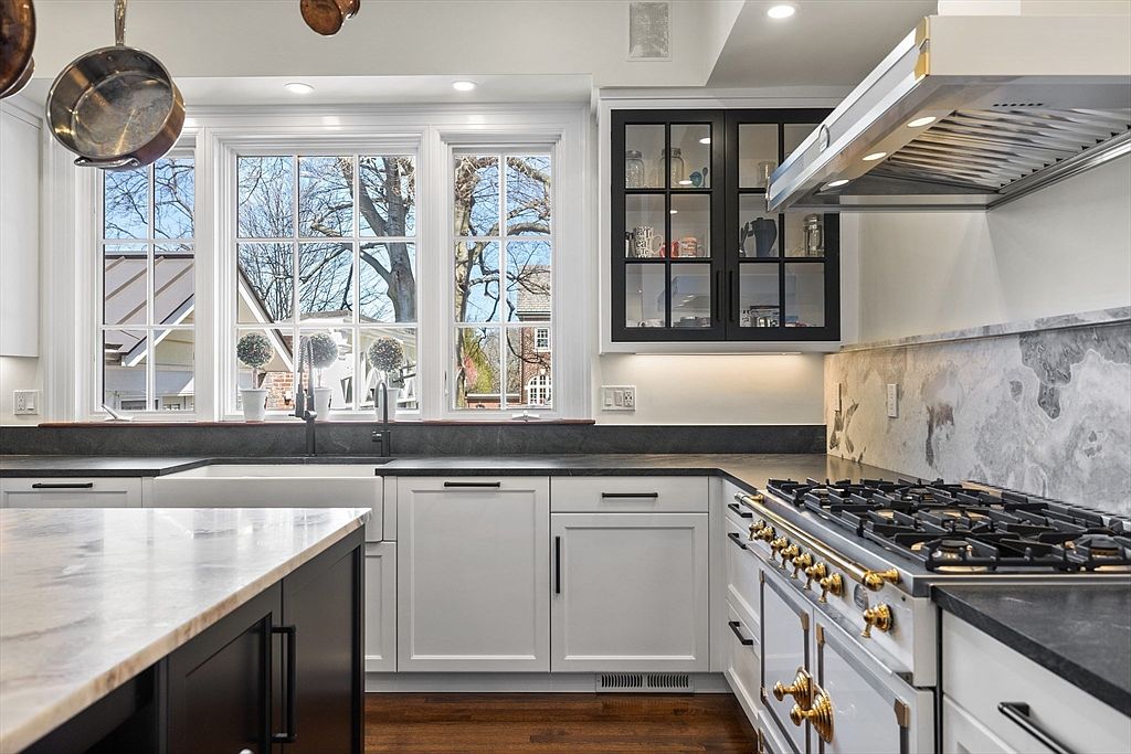 This elegant kitchen features a blend of white and dark cabinetry, complemented by dark stone countertops and a striking stone backsplash behind a professional-style range. A large window above the farmhouse sink provides ample natural light and a view of the outdoors, while a glass-front cabinet adds a touch of sophistication. The perspective captures the functional layout, highlighting the high-end appliances and the warm wood flooring.